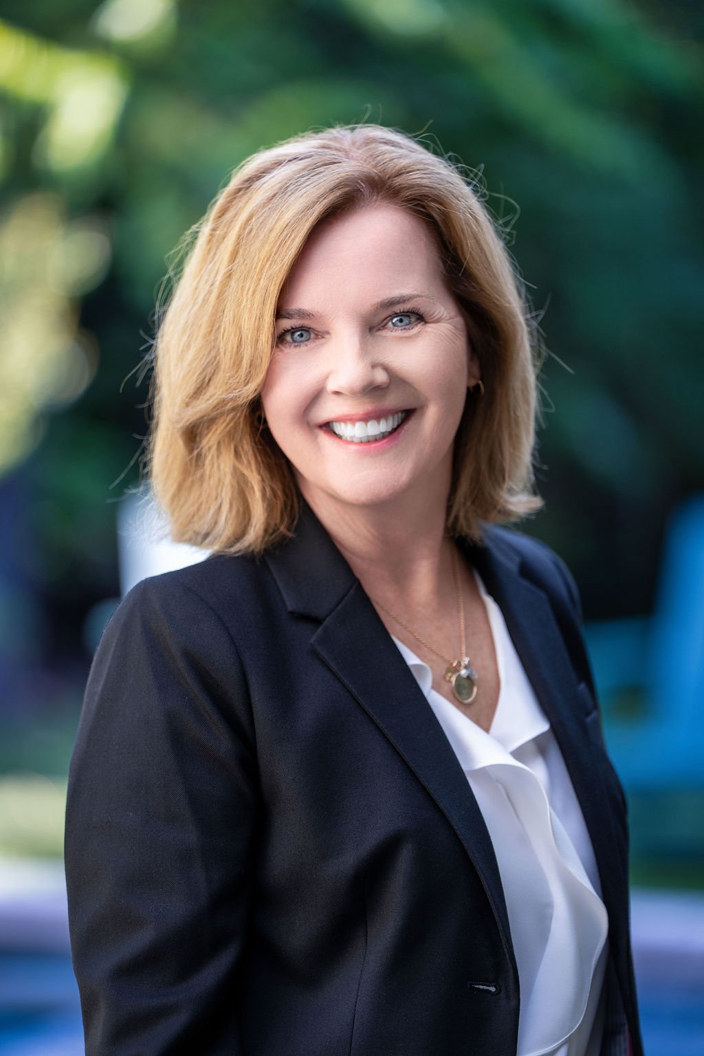 A smiling woman with shoulder-length blonde hair, wearing a black blazer over a white blouse, standing outdoors with blurred green foliage in the background.