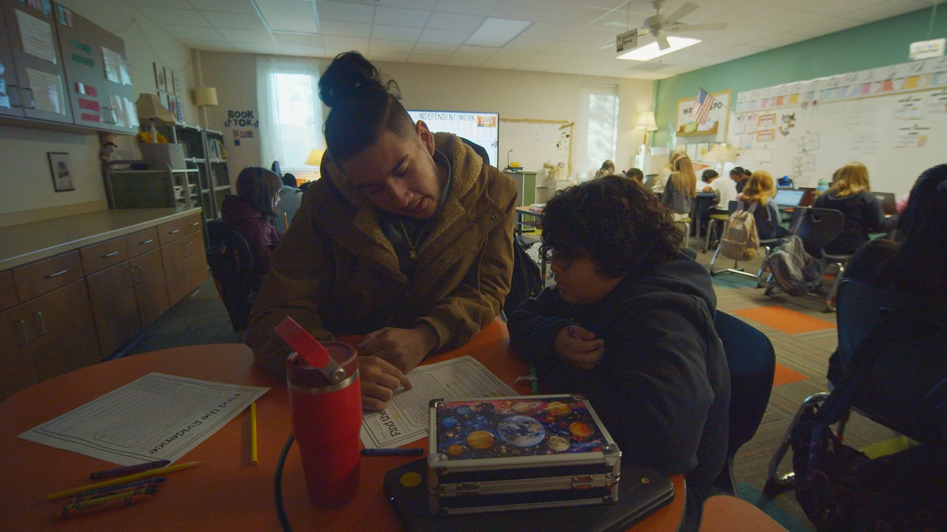 A teacher helps a student with her work in a classroom filled with students working at tables and on laptops, with educational posters on the walls.