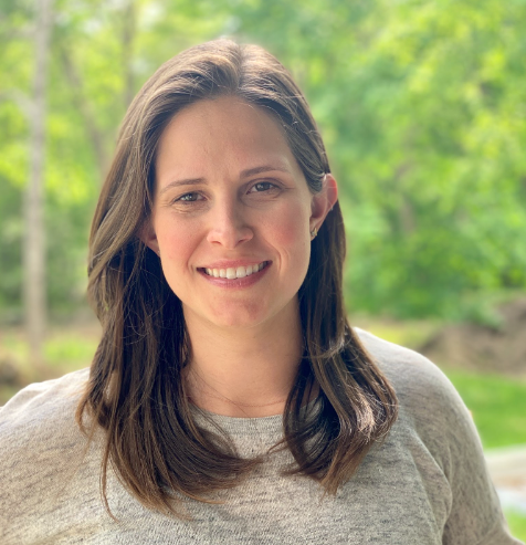 A young woman with brown hair smiling outdoors with green trees in the background.