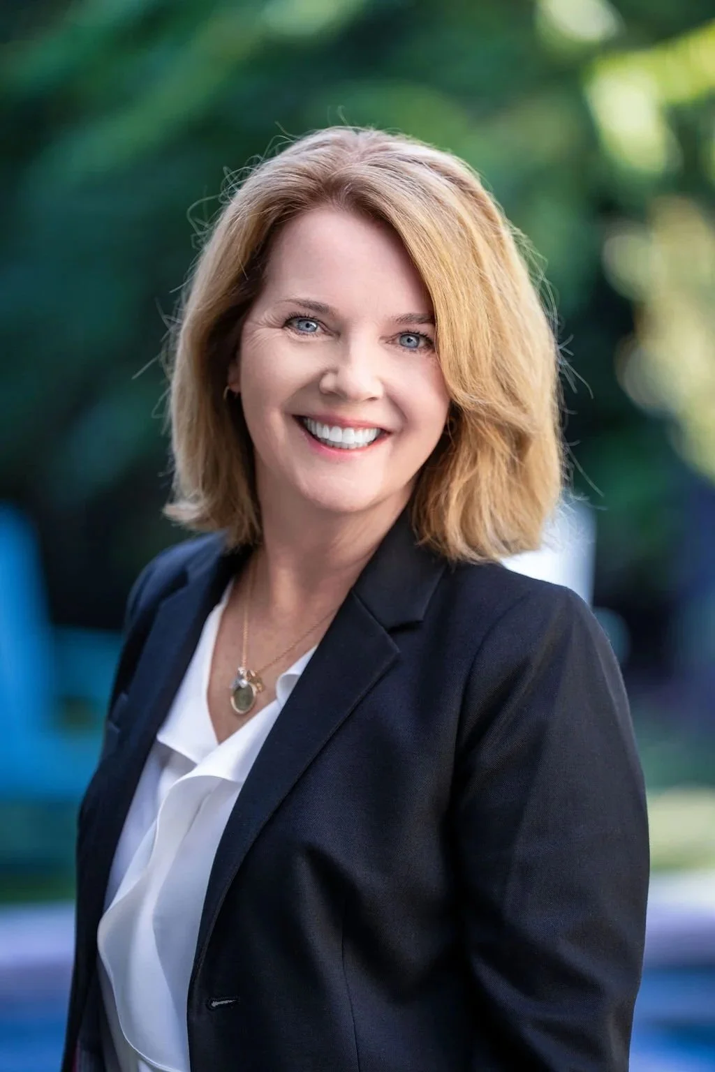 A smiling woman with shoulder-length blonde hair, wearing a black blazer over a white blouse, standing outdoors with blurred green foliage in the background.
