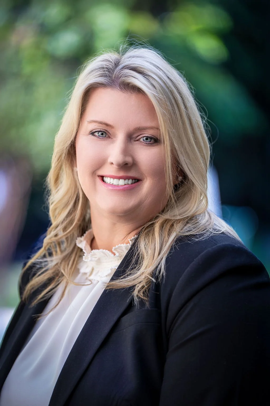 A blonde woman smiling, dressed in a black blazer and white blouse, outdoors with blurred green foliage in the background.