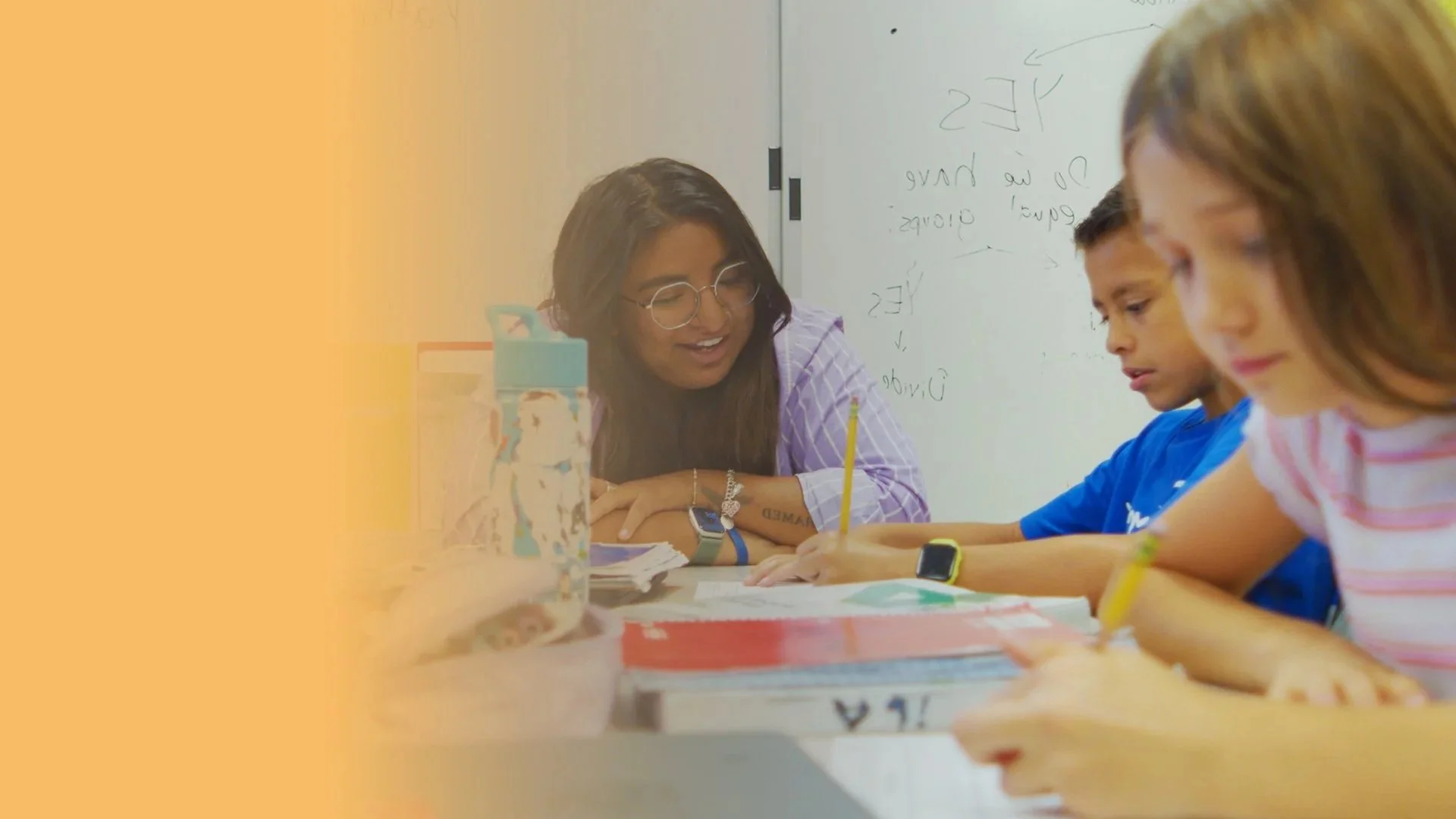 A teacher sitting at a desk with three students, engaging with them during a classroom activity. The students are writing with yellow pencils on paper, and the teacher is smiling and looking at them. There is a whiteboard with writing behind them.