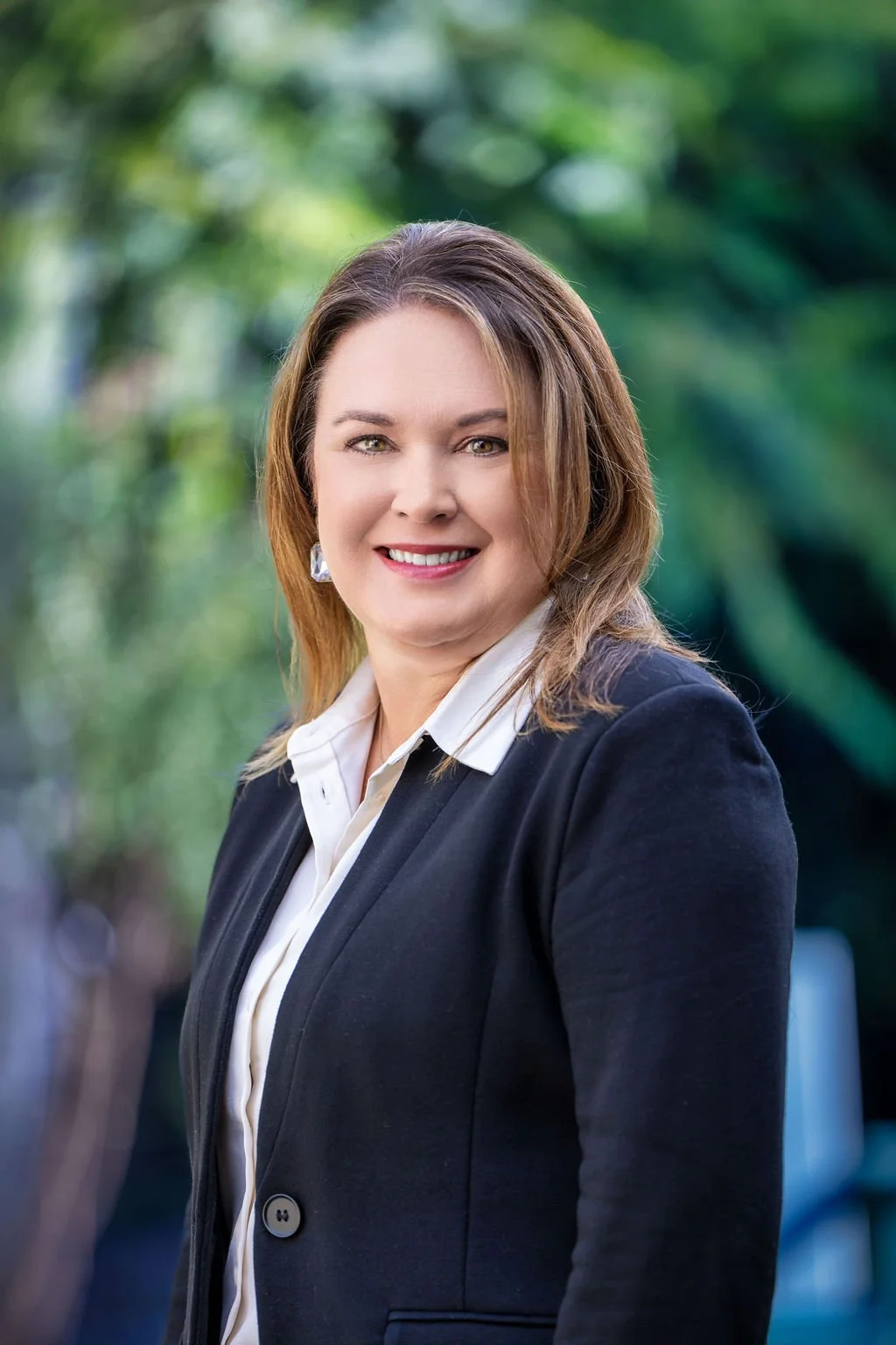 A woman with shoulder-length brown hair, wearing a black blazer over a white shirt, smiling outdoors with blurred green foliage background.