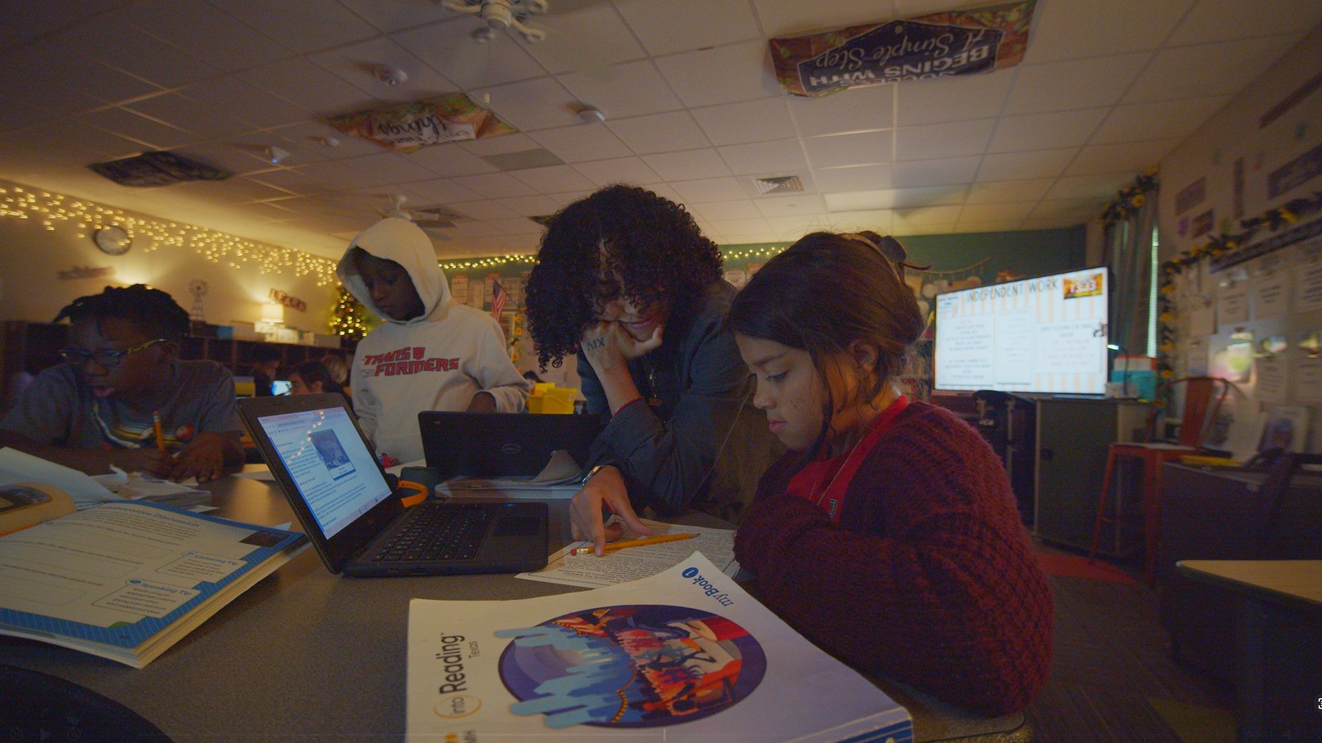 A teacher assisting a young girl with her schoolwork in a classroom, with other students working at tables, a large screen displaying a presentation, and classroom decorations in the background.