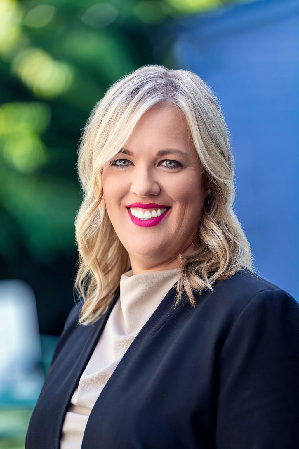 A woman with shoulder-length blonde hair, wearing a black blazer and a beige top, smiling with pink lipstick against a blurred outdoor background with green foliage and blue sky.