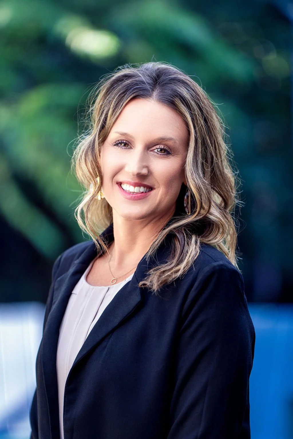 A smiling woman with wavy, shoulder-length hair in an outdoor setting with trees and greenery in the background.