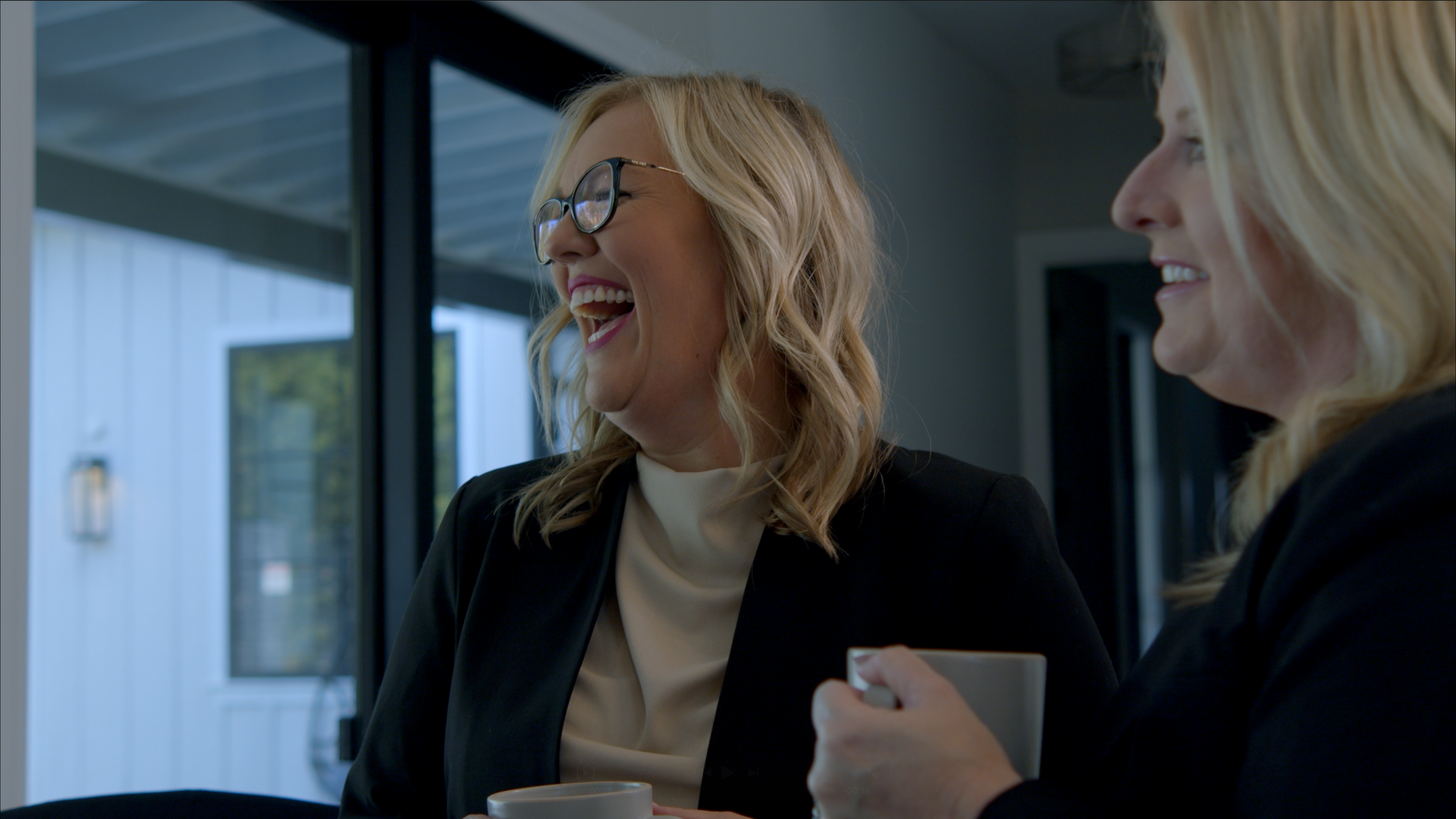 Two women with blonde hair and black blazers are smiling and laughing while holding coffee mugs indoors.