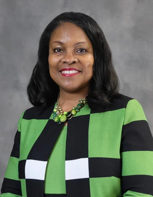 A woman with shoulder-length black hair wearing a green and black checkered blazer, a green top, and a green beaded necklace, standing against a gray background, smiling at the camera.