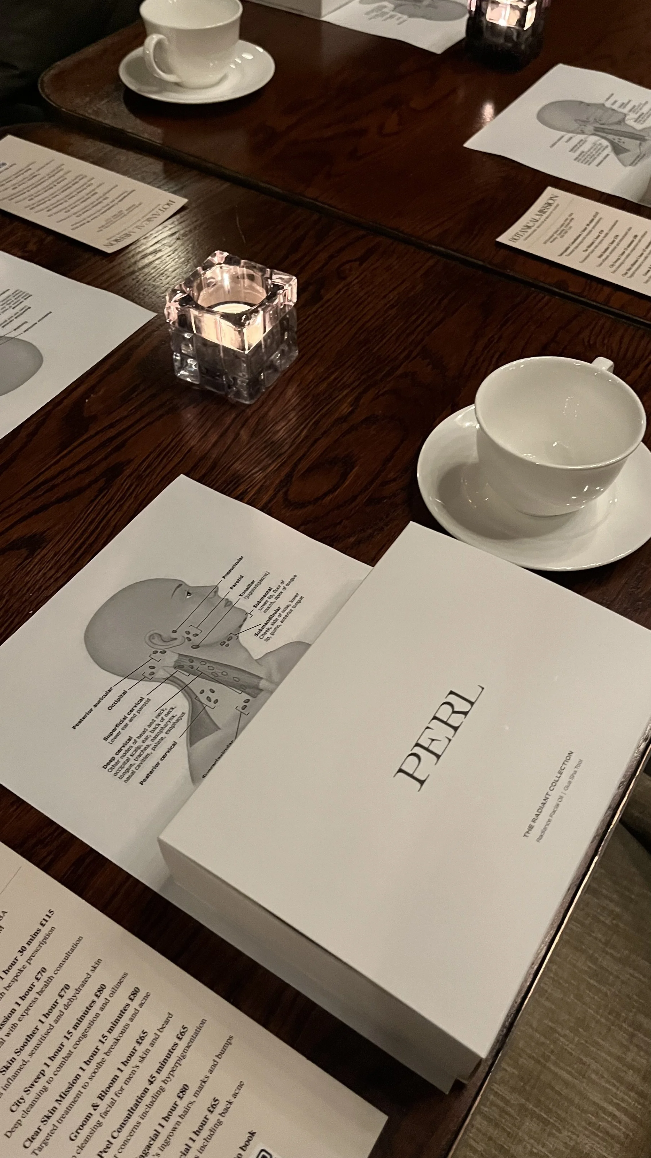A restaurant table setting with a white box labeled 'PEAR', a printed diagram of an ear and neck, a white teacup on a saucer, a small lit candle in a clear square holder, and printed menus.