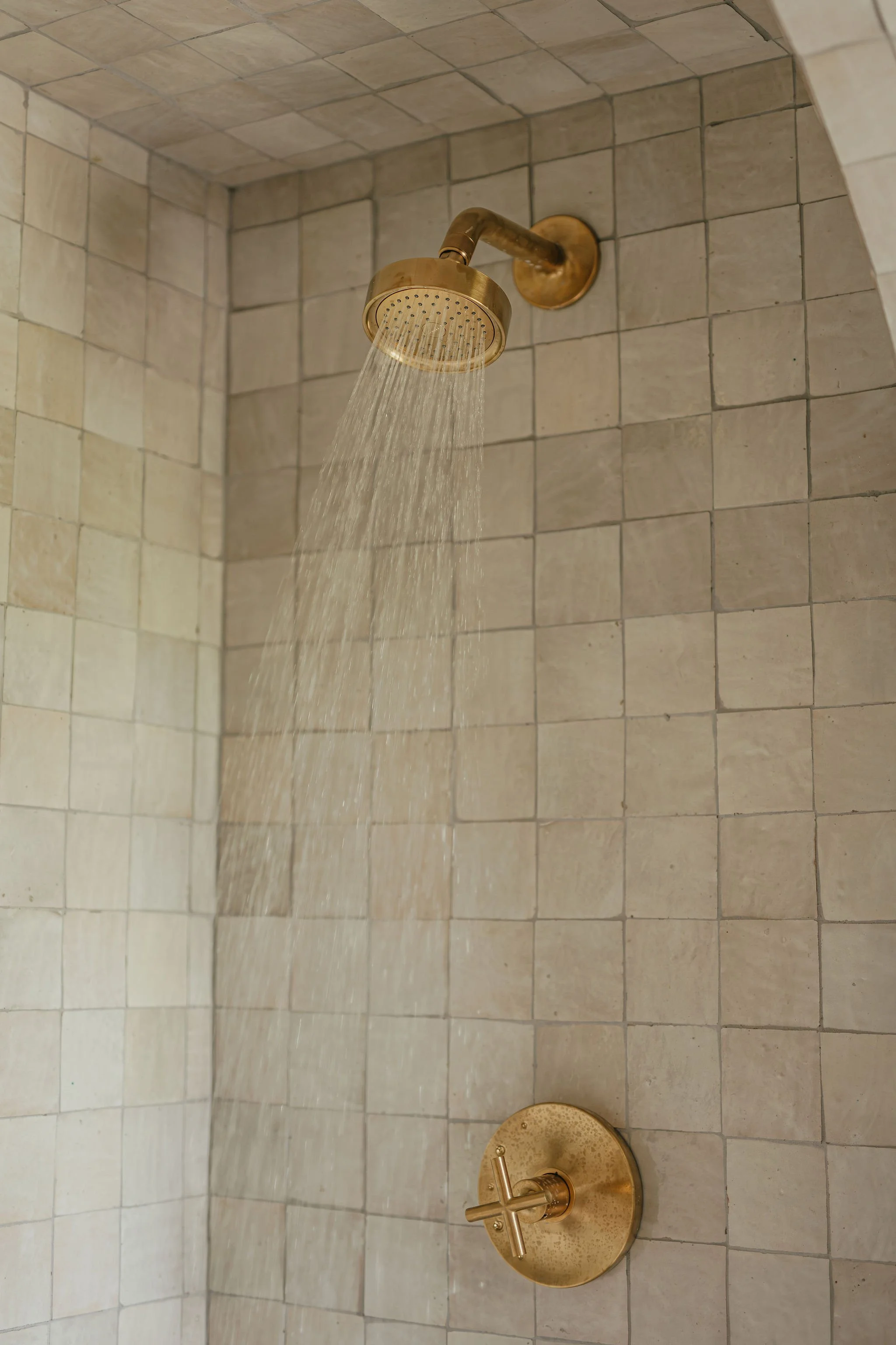 Brass showerhead with water running, mounted on tiled wall with beige tiles.
