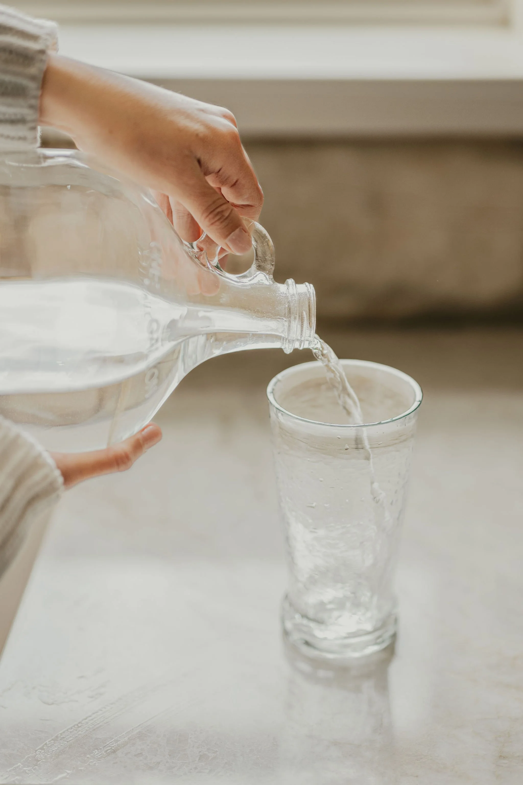 Person pouring water from a glass pitcher into a tall glass on a neutral-colored surface.