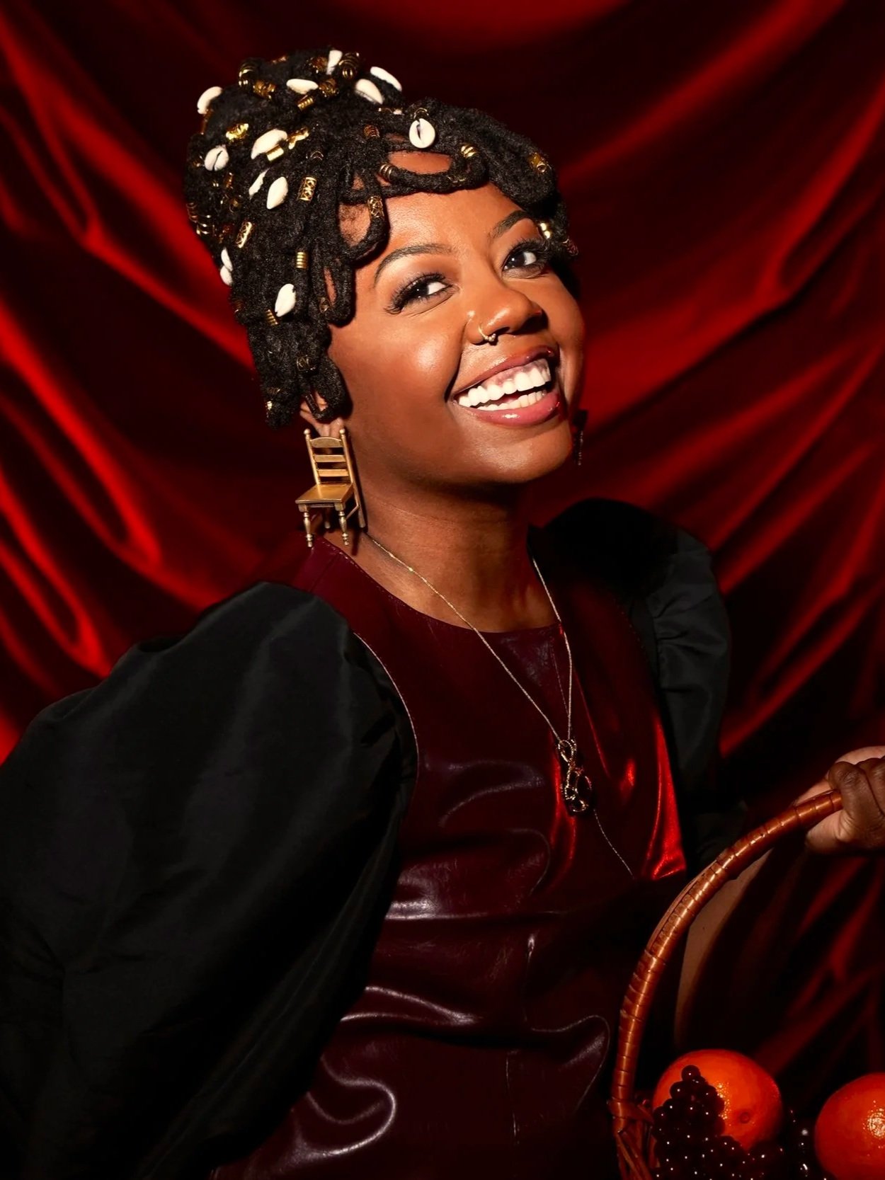 A woman with styled dreadlocks adorned with white shells and gold accents, smiling brightly, wearing large earring and necklaces, holding a basket of fruit against a red velvet backdrop.