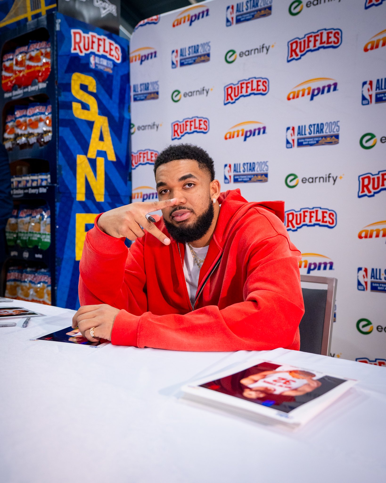 A man in a red hoodie sitting at a table making a peace sign with his right hand, at a sports-themed autograph signing event with a backdrop featuring NBA All-Star 2025 and Ruffles logos.