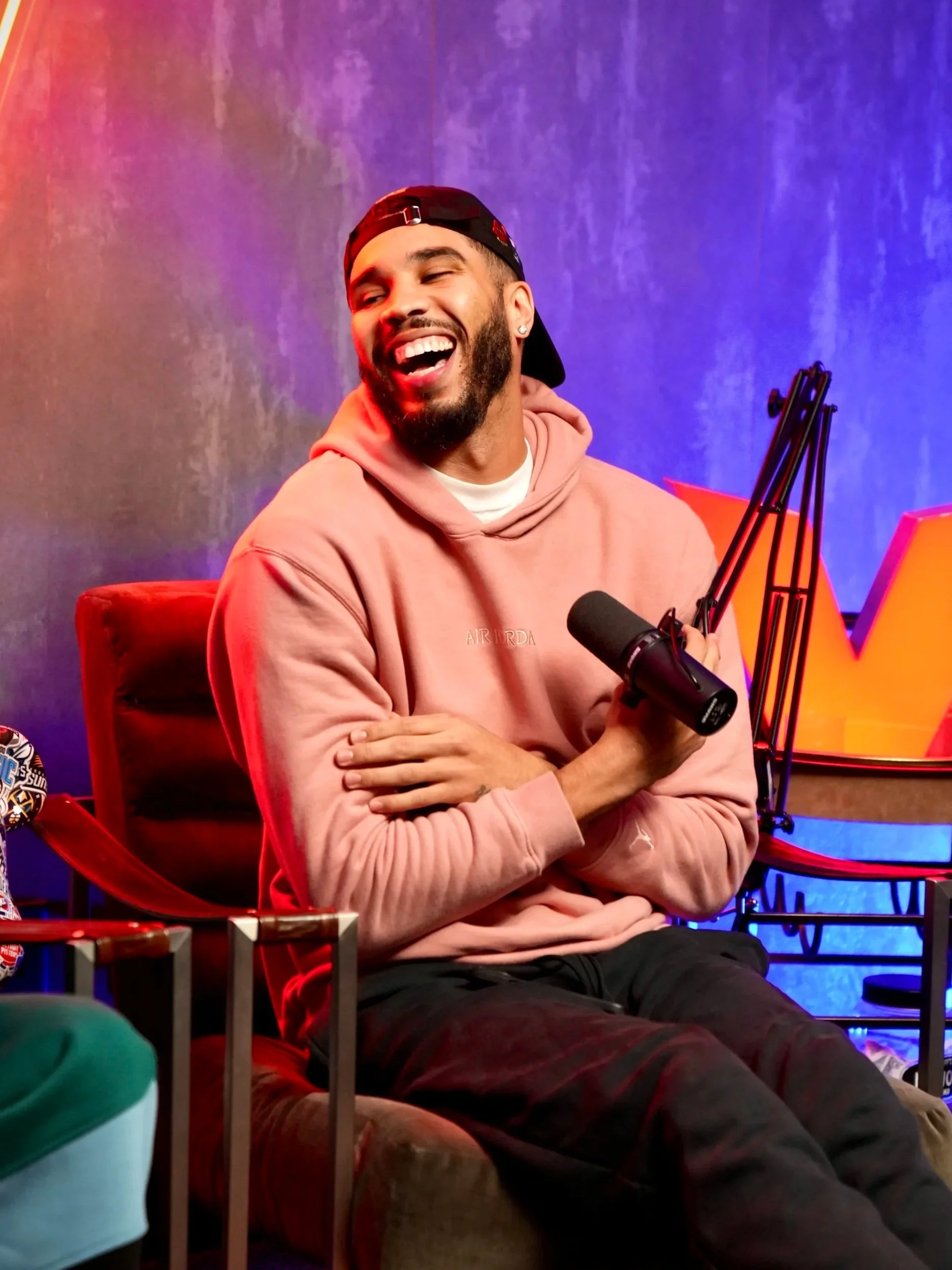 A man wearing a pink hoodie and a backwards cap, smiling and laughing while holding a microphone in a studio setting with purple and blue lighting. Jayson Tatum