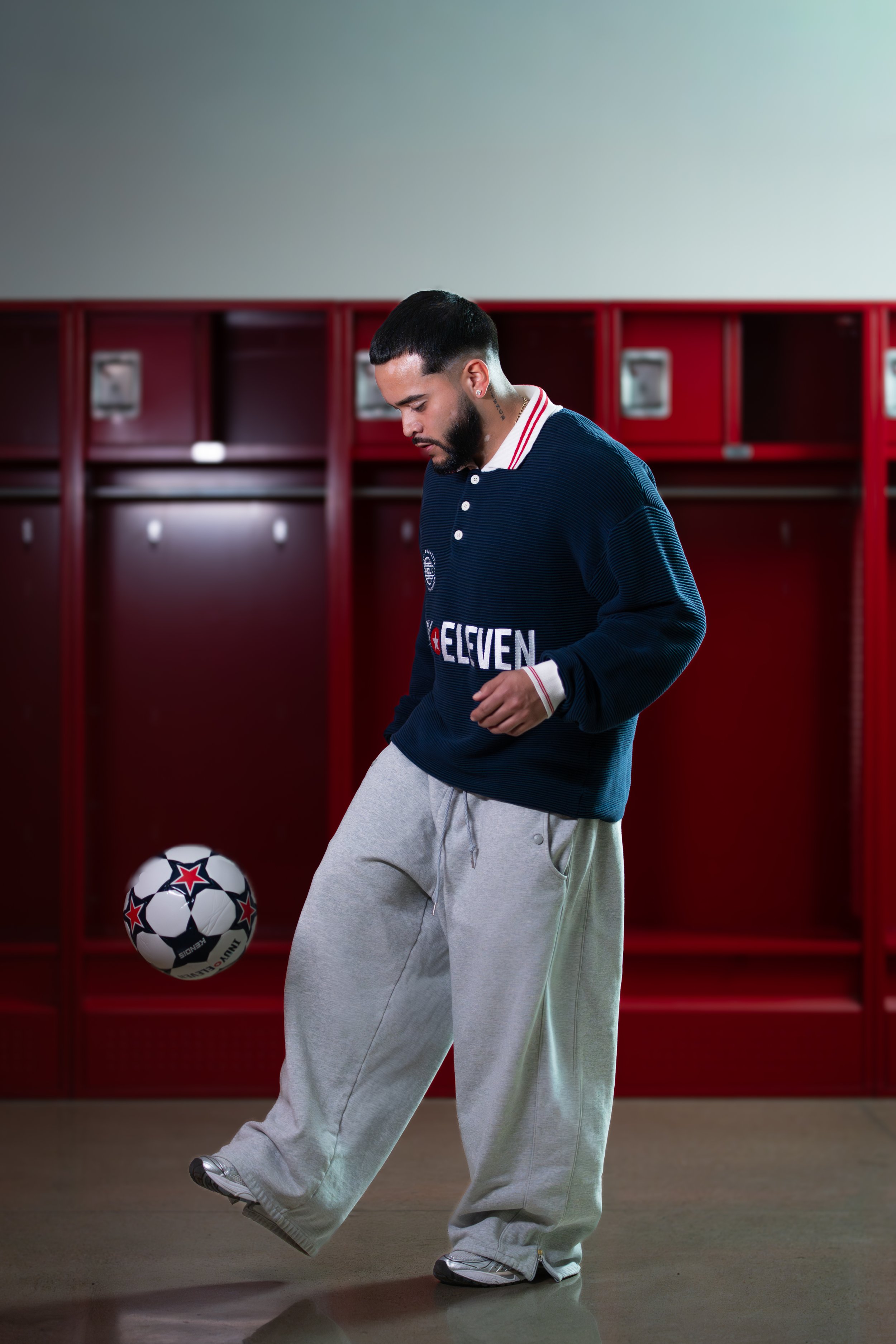 A young man with a beard and tattoos on his neck and arm, wearing a navy blue soccer jersey with 'Eleven' written on it, gray sweatpants, and white shoes, is in a locker room kicking a soccer ball with red, black, and white patterns.