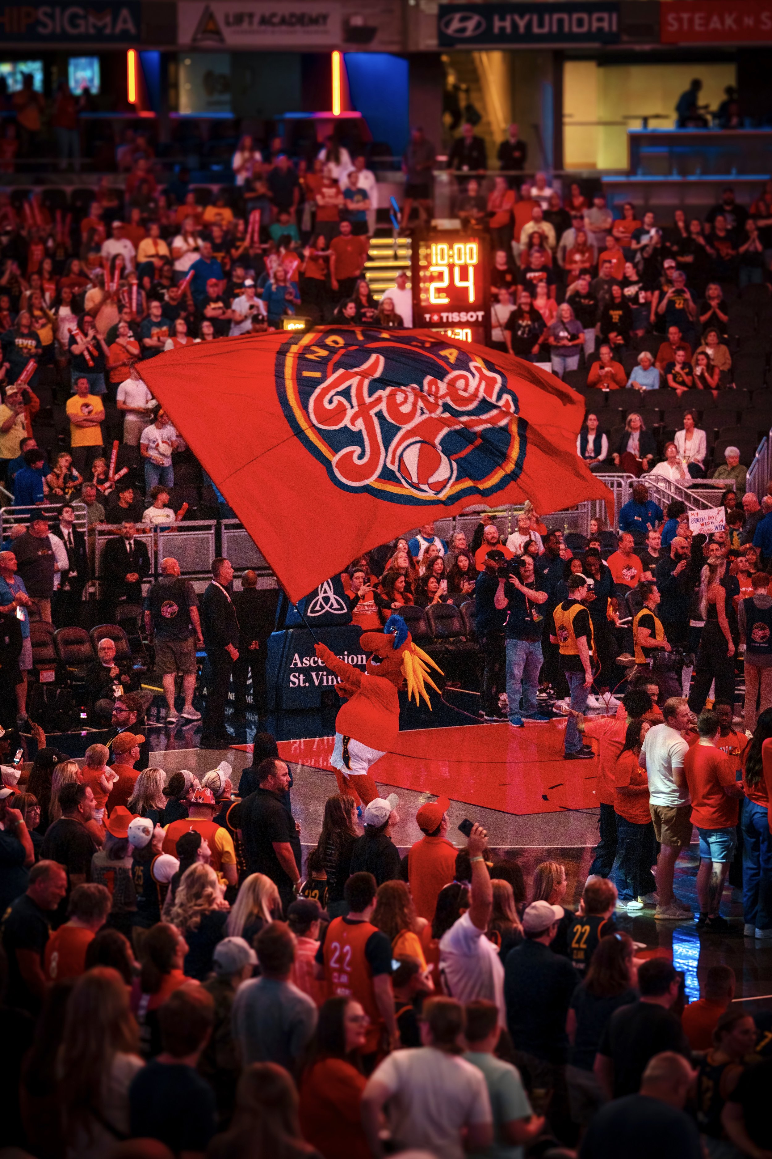 Crowd watching a basketball game, with a mascot waving a large red flag with the Fenerbahçe logo, inside an indoor sports arena.
