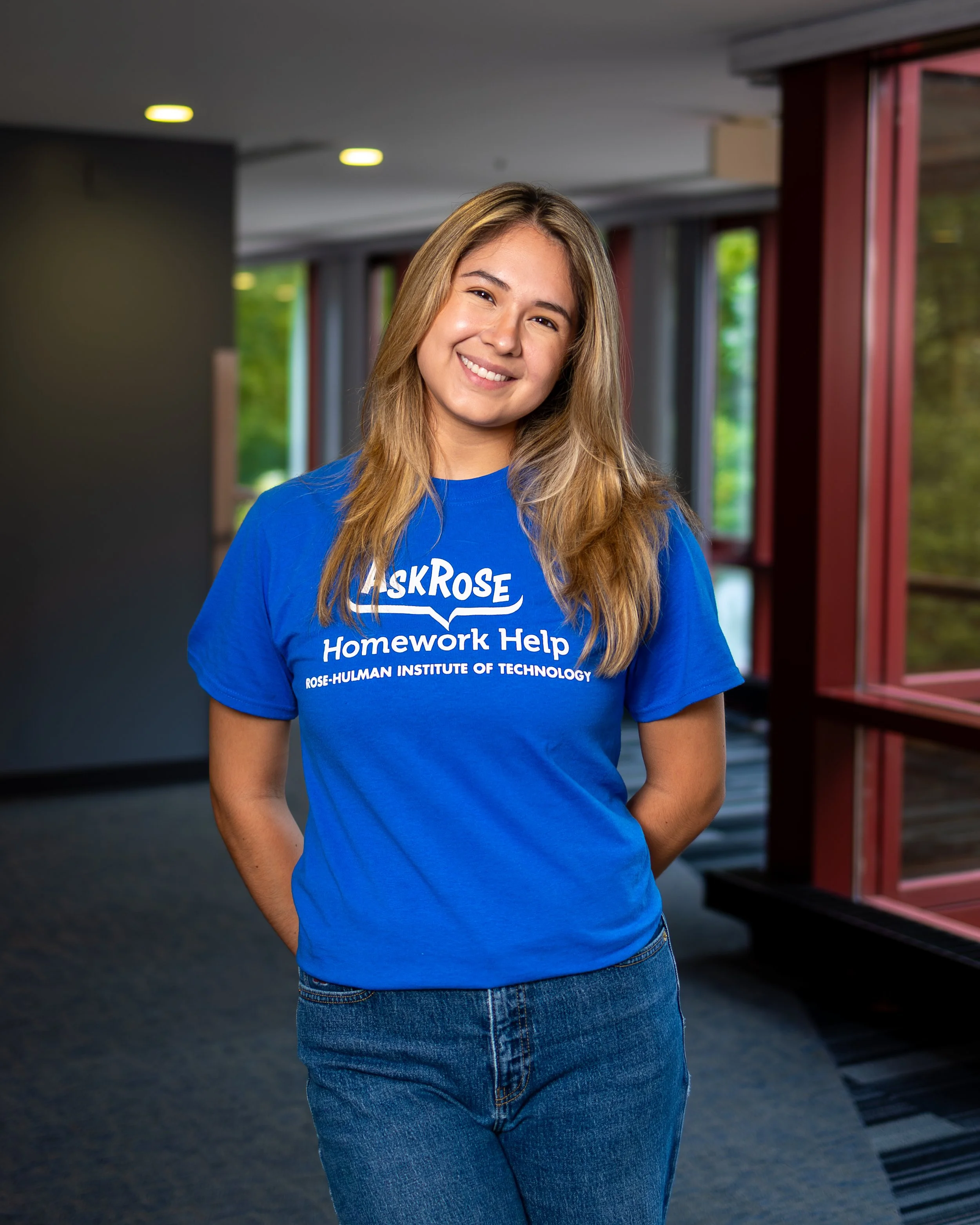A young woman with long blonde hair smiling, wearing a blue T-shirt with white text that reads 'Ask Rose Homework Help' from Rose-Hulman Institute of Technology, standing in an indoor space with large windows showing green trees outside.