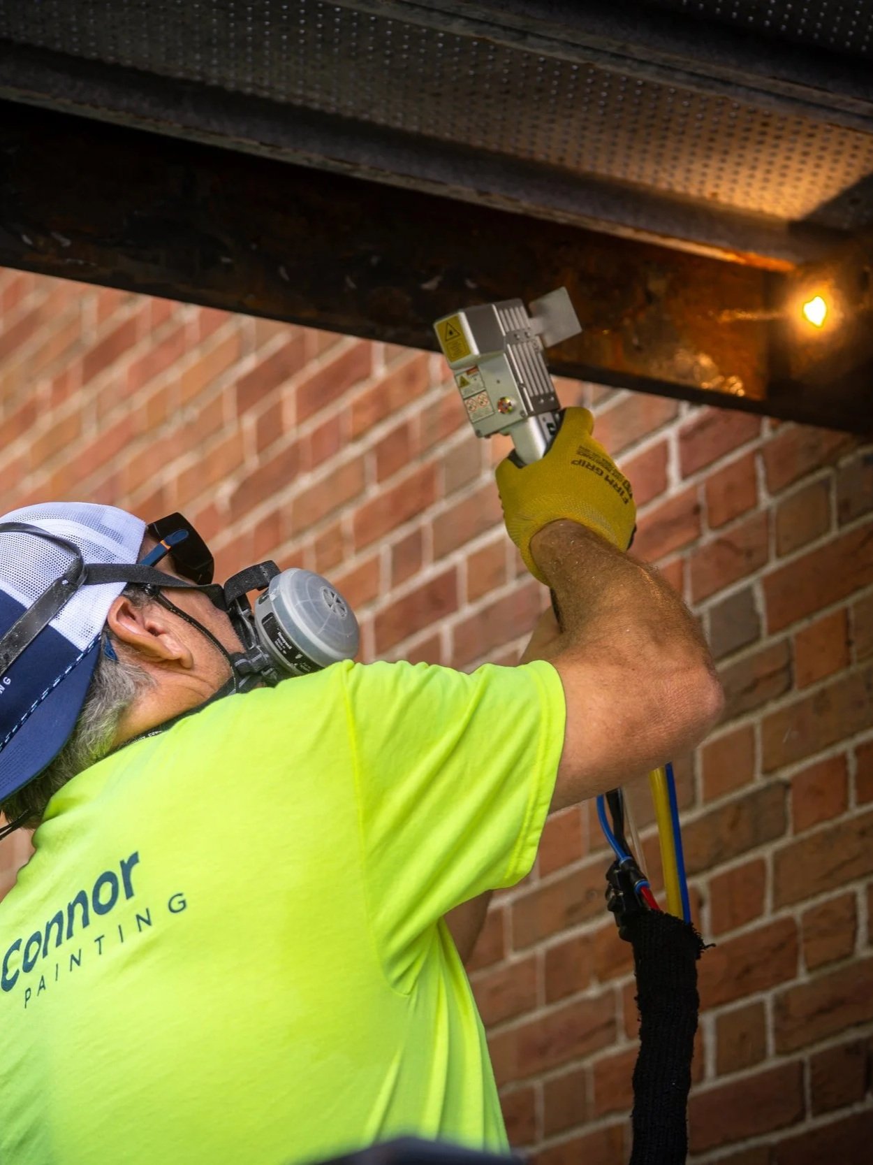 A worker in a yellow shirt, gloves, and a baseball cap with a respirator mask is using a power tool on a metal beam under a building structure, with a brick wall in the background.