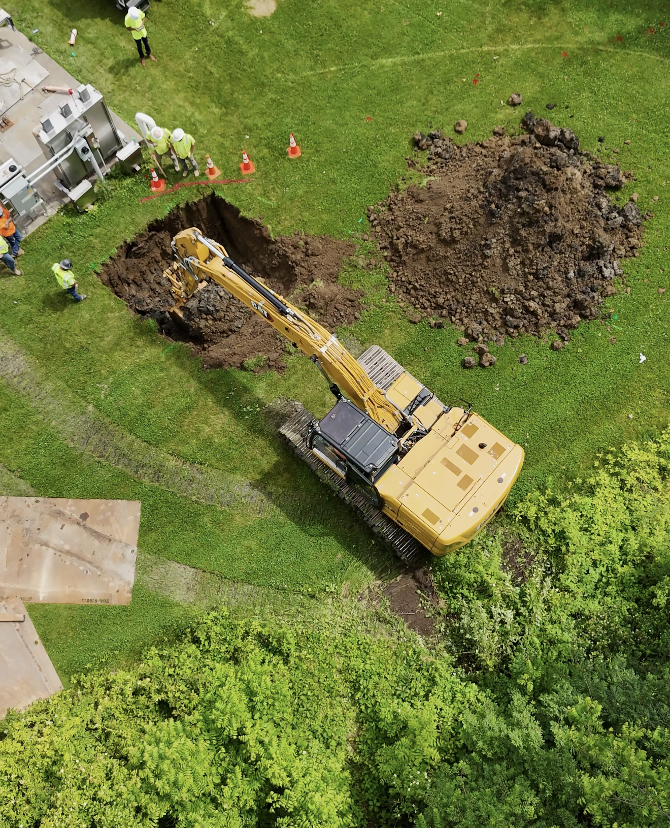 An aerial view shows a construction site with a yellow excavator digging a trench in a grassy area, surrounded by workers wearing safety helmets and vests, some standing near traffic cones and equipment.