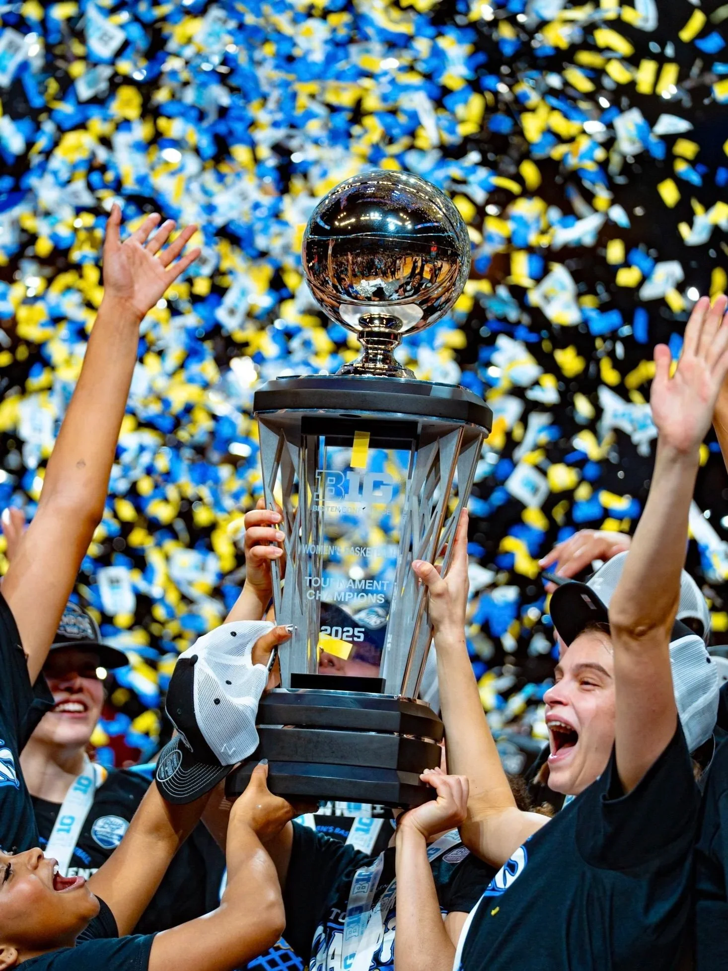The women's basketball team celebrating with a trophy amidst blue, white, and yellow confetti after winning a tournament in 2025.