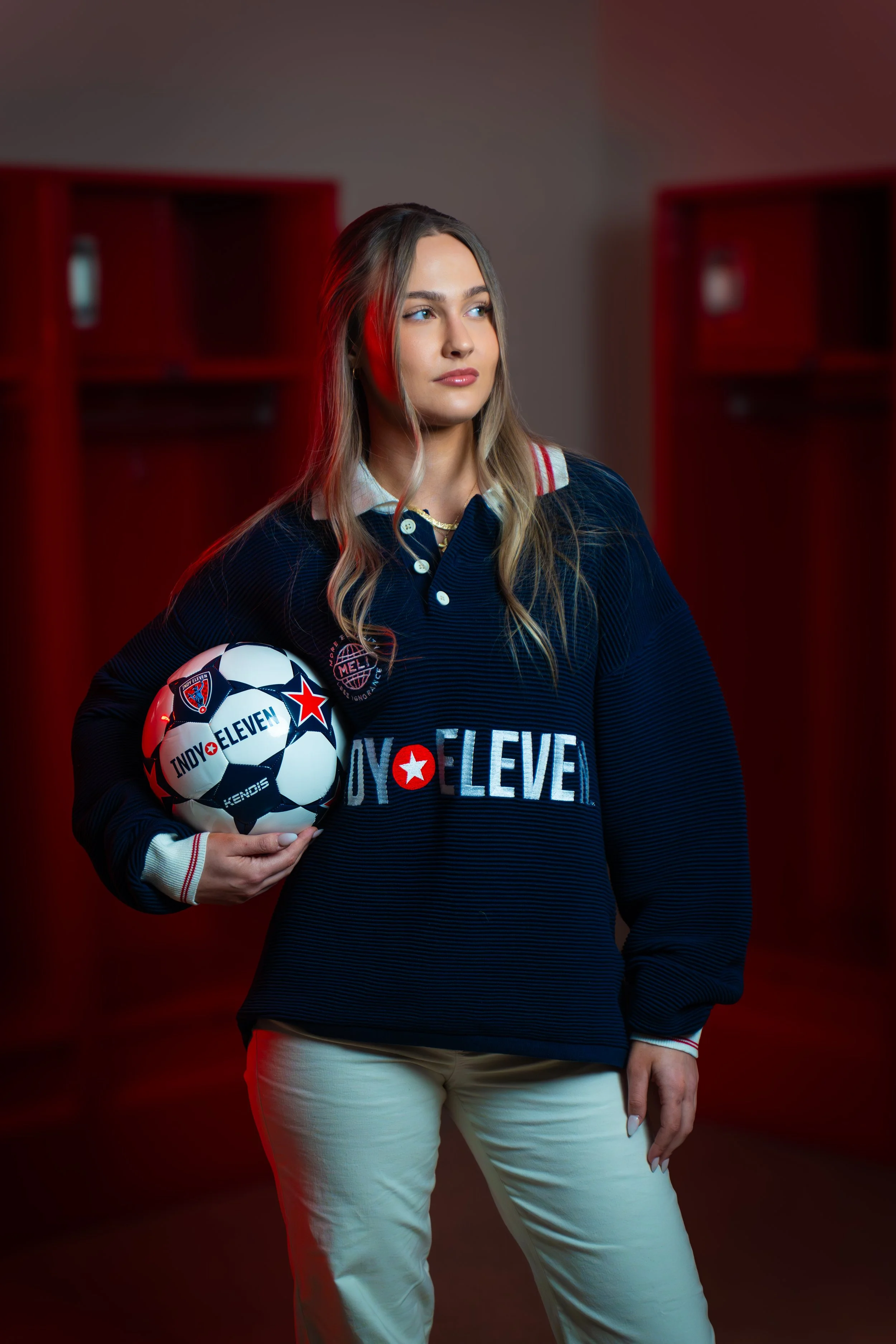 A woman holding a soccer ball in a locker room, wearing a navy blue sweater with logos, blonde hair, and beige pants.