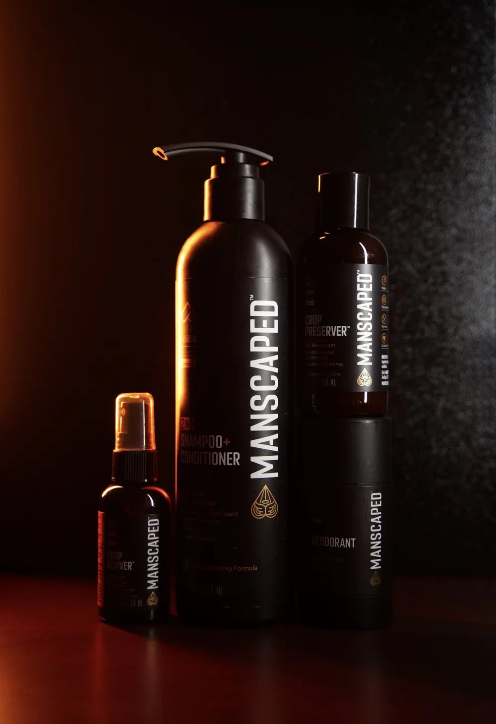 Collection of black bottles with white and gold labels labeled 'MANSCAPED', including shampoo, conditioner, deodorant and pre-shave product, against a dark background with orange and yellow lighting accents.