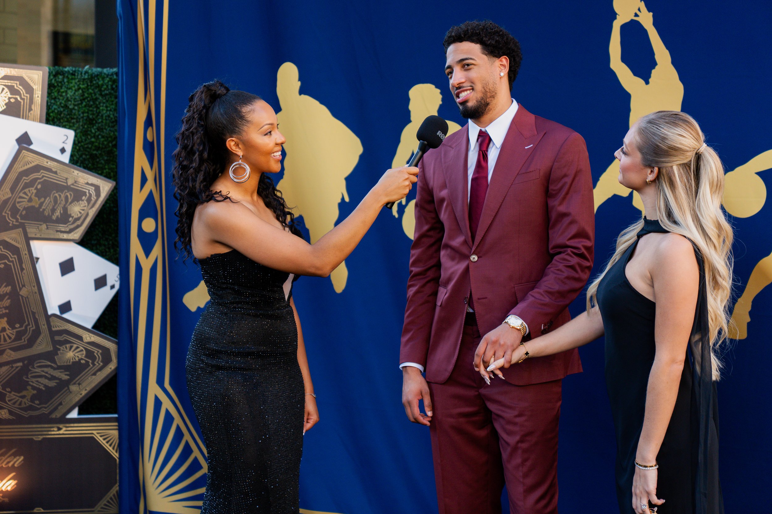 A woman interviewing a man and woman on a red carpet at an event, with a blue backdrop featuring gold silhouettes of performers.