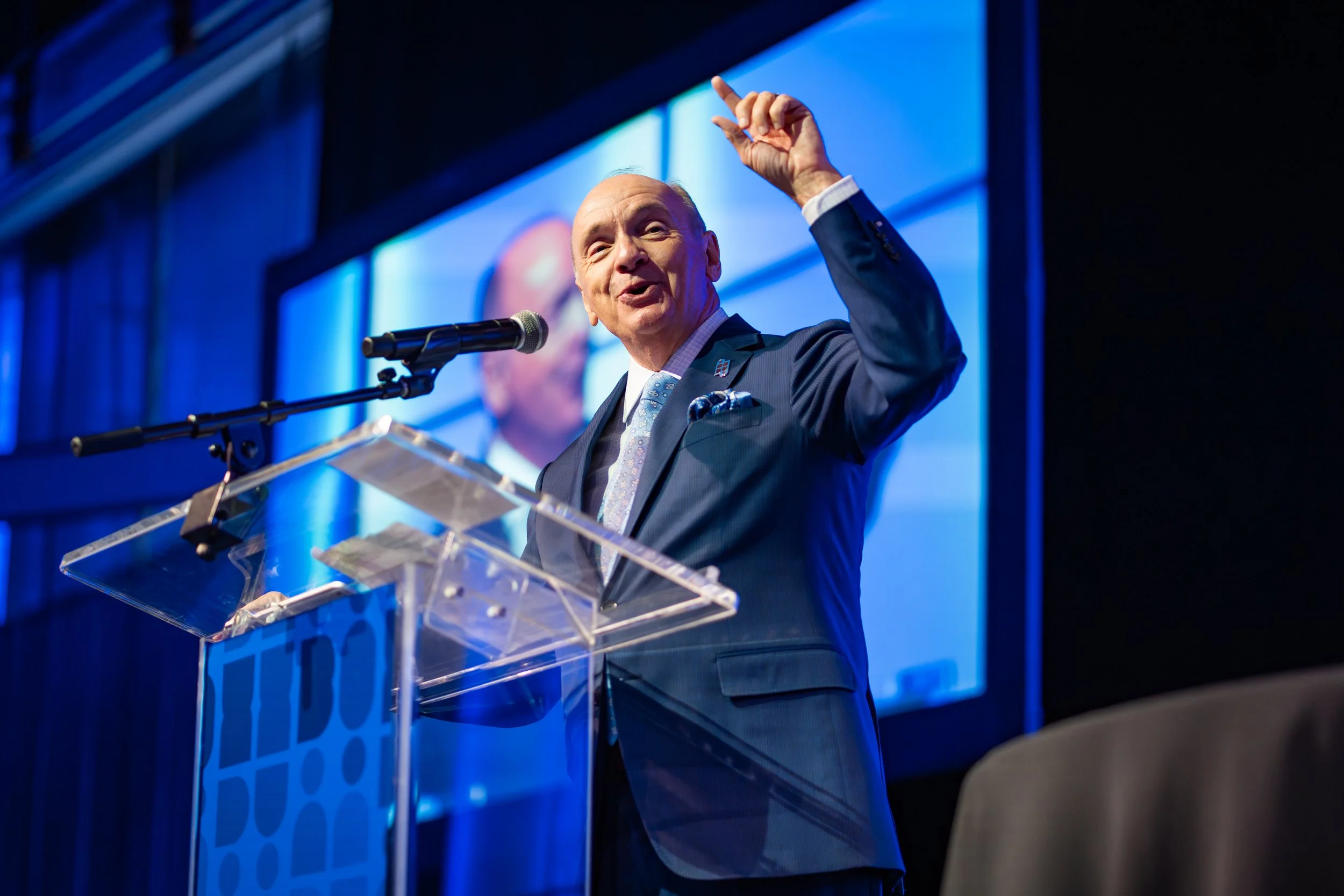 A man in a dark suit and a tie speaking at a podium with a microphone, gesturing with his hand, with a large screen displaying his image behind him.
