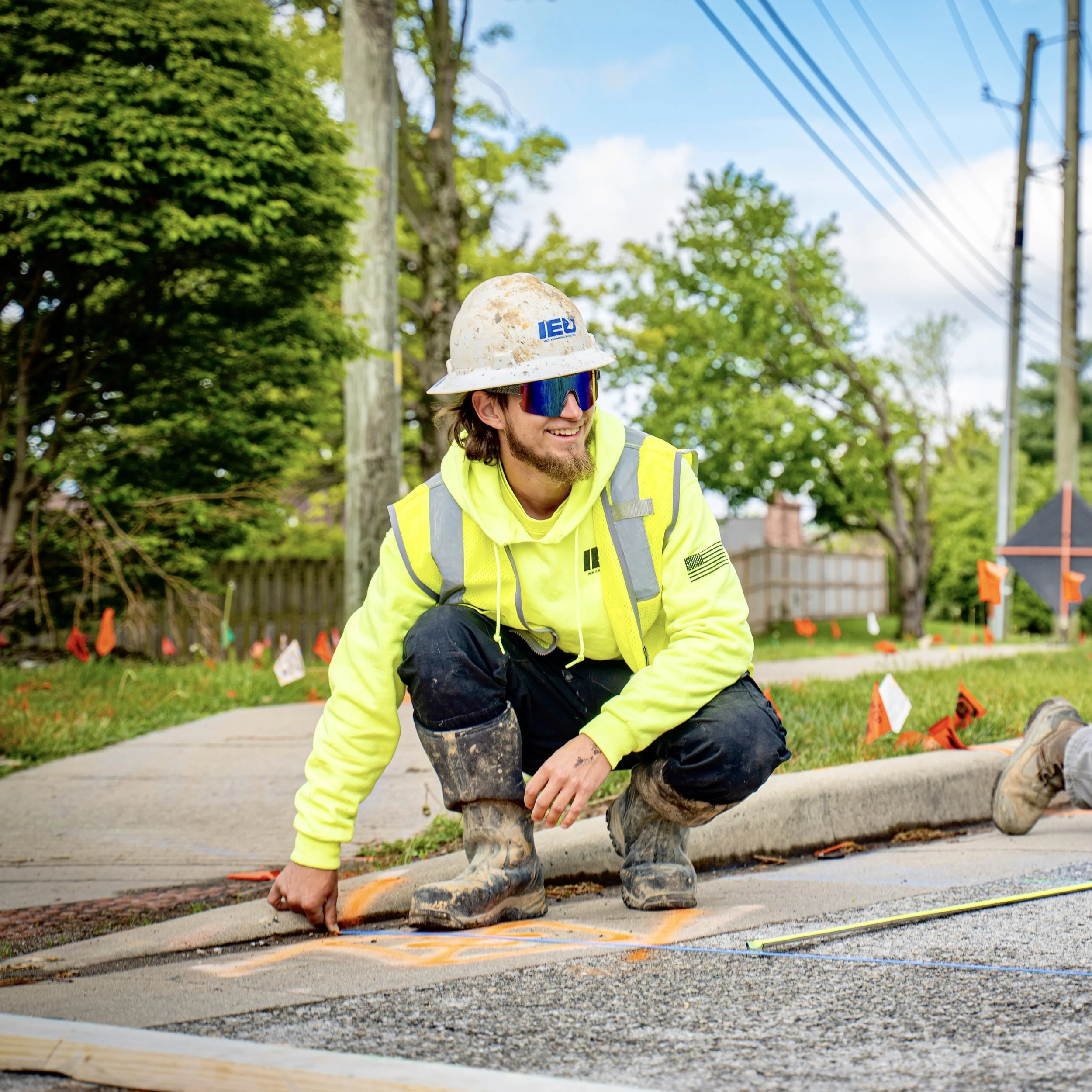 Construction worker in a yellow safety vest and hard hat crouching on a sidewalk during daytime, working on pavement with trees and power lines in the background.