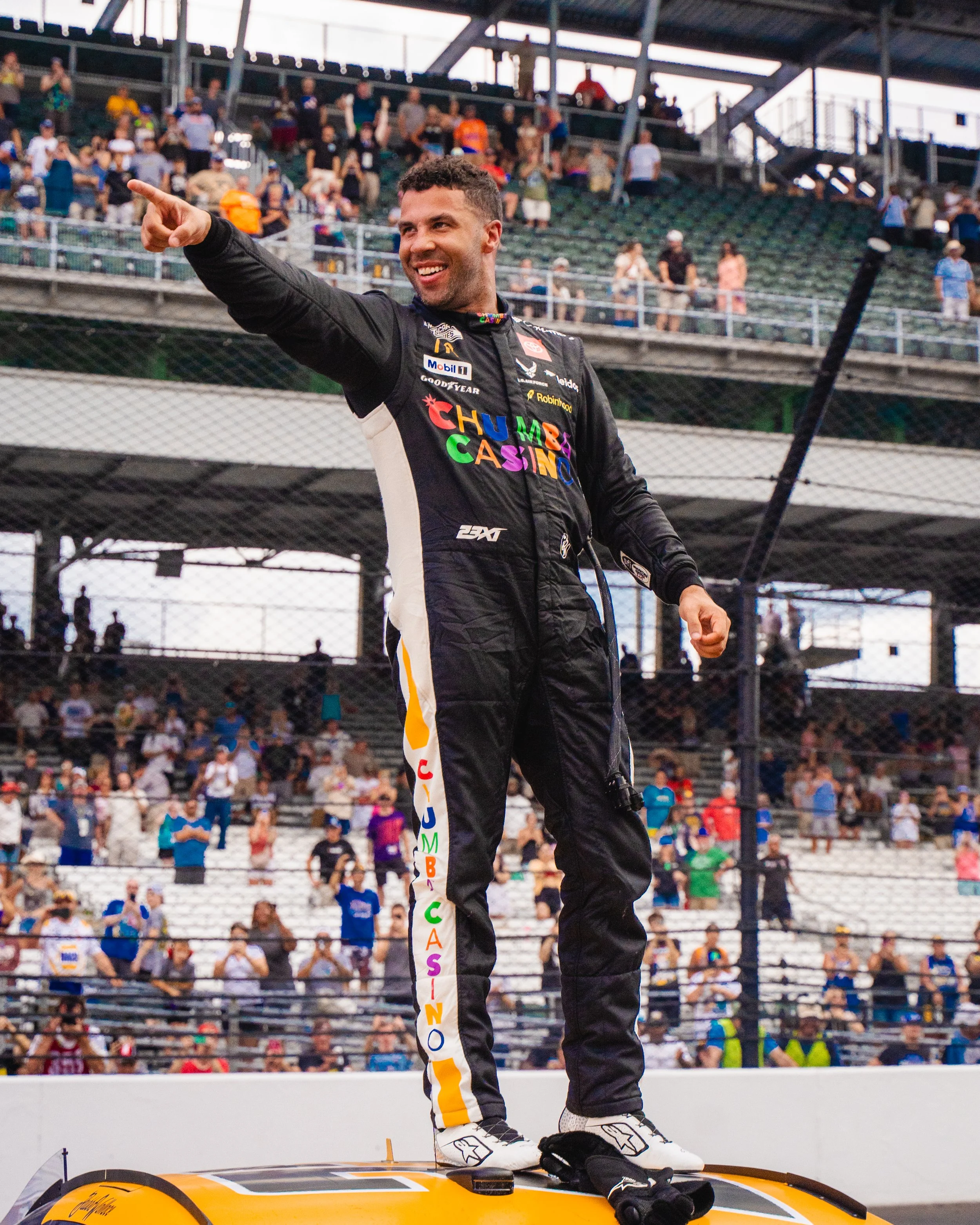 Race car driver in a black racing suit with colorful logos, smiling and pointing outward on the victory podium with a crowd in the background.