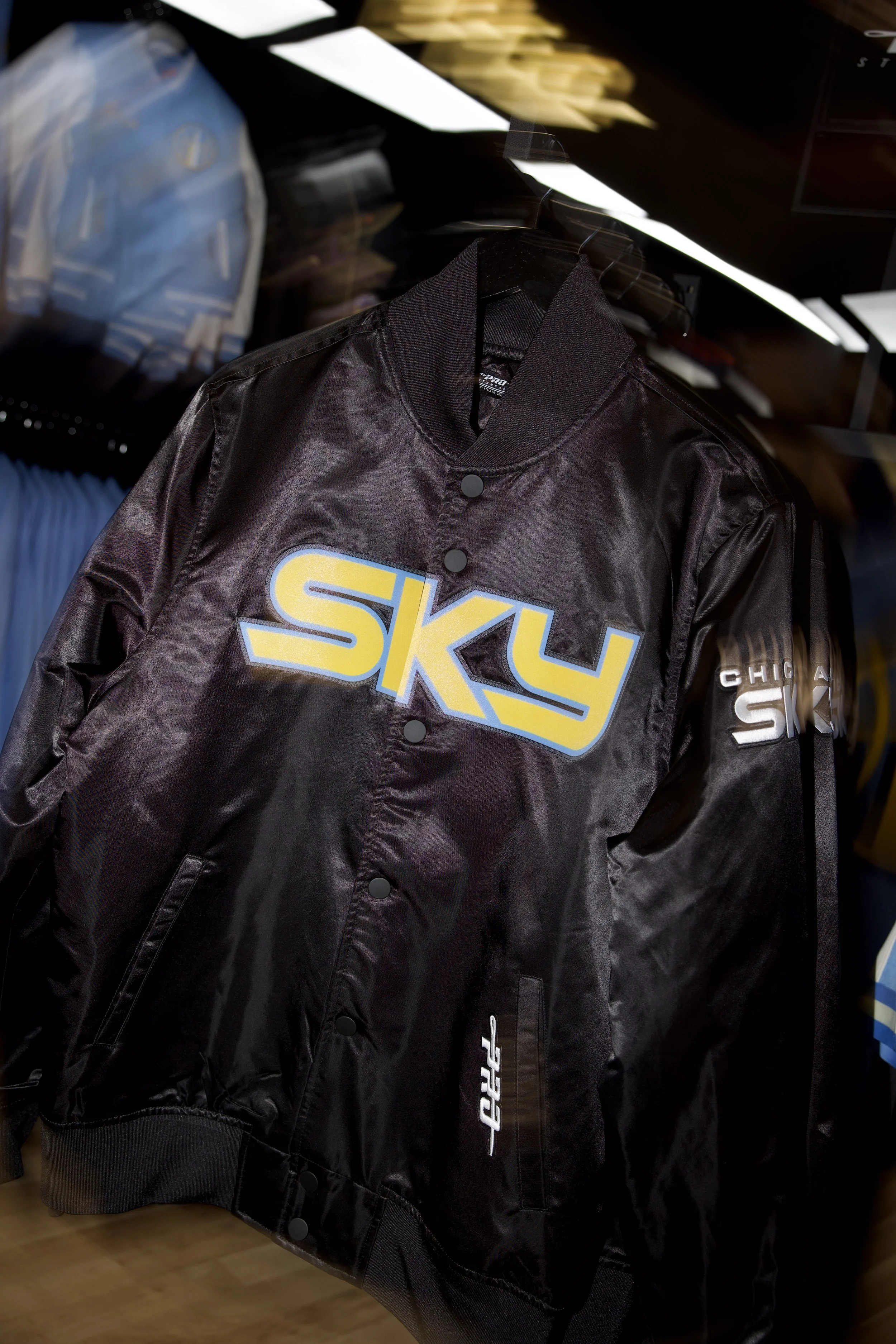 Black bomber jacket with large yellow and blue 'SKY' logo on the front, hanging on a black hanger in a store.