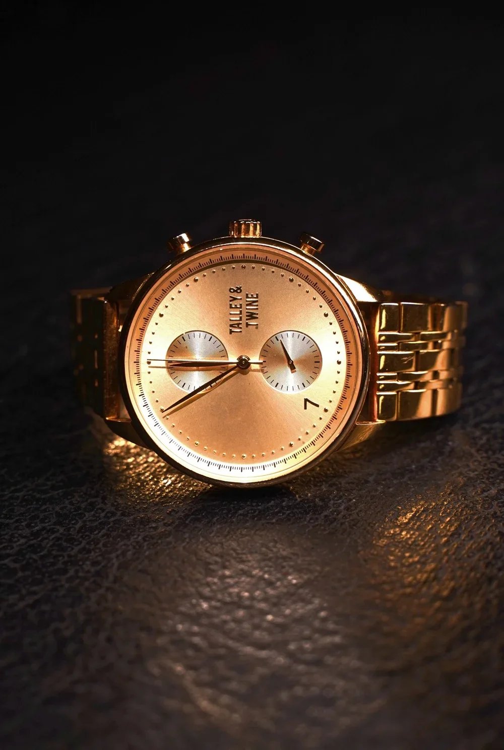 A gold wristwatch with a round face, two smaller dials, and a metallic band, placed on a textured surface with soft lighting.
