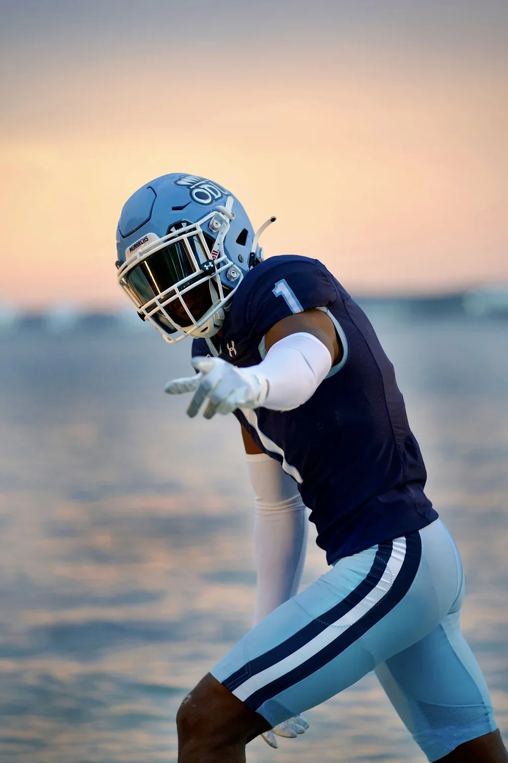 A football player in uniform and helmet on the beach during sunset, pointing towards the camera.