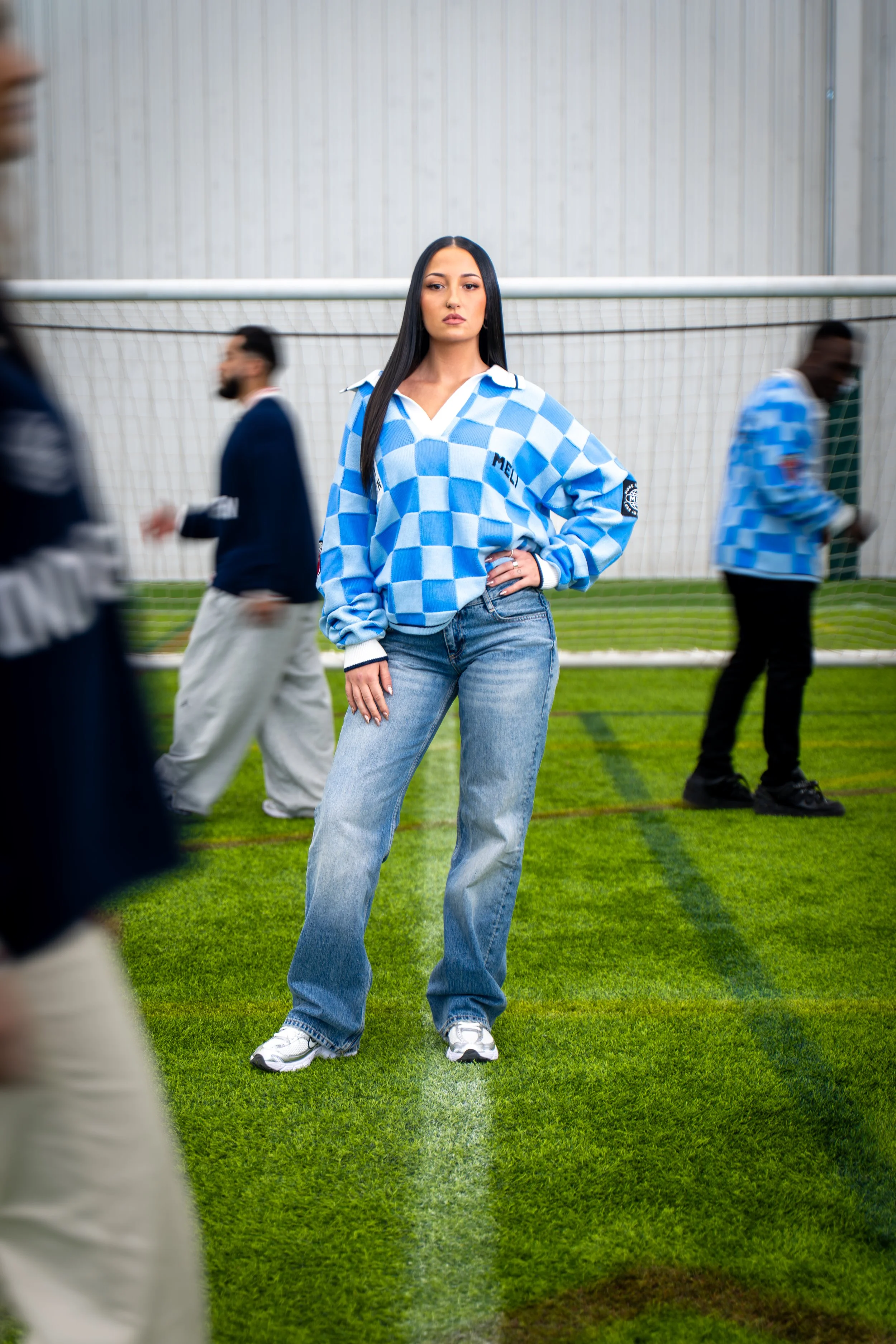 Young woman with long black hair standing on a soccer field, wearing a blue checkered sweater and jeans, surrounded by blurred people.