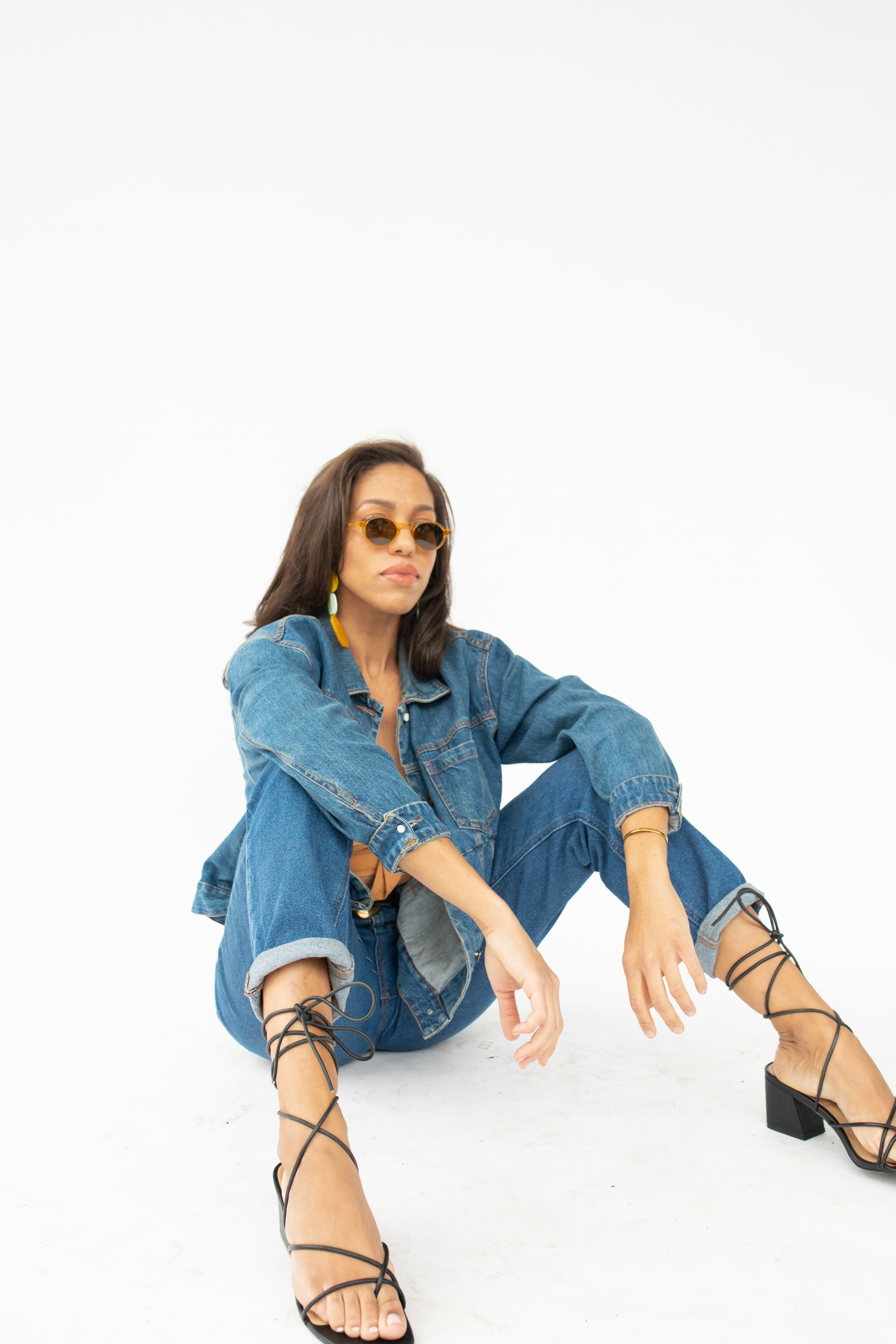 Woman wearing sunglasses, denim jacket and jeans, black strappy heels, sitting on white floor against white background.