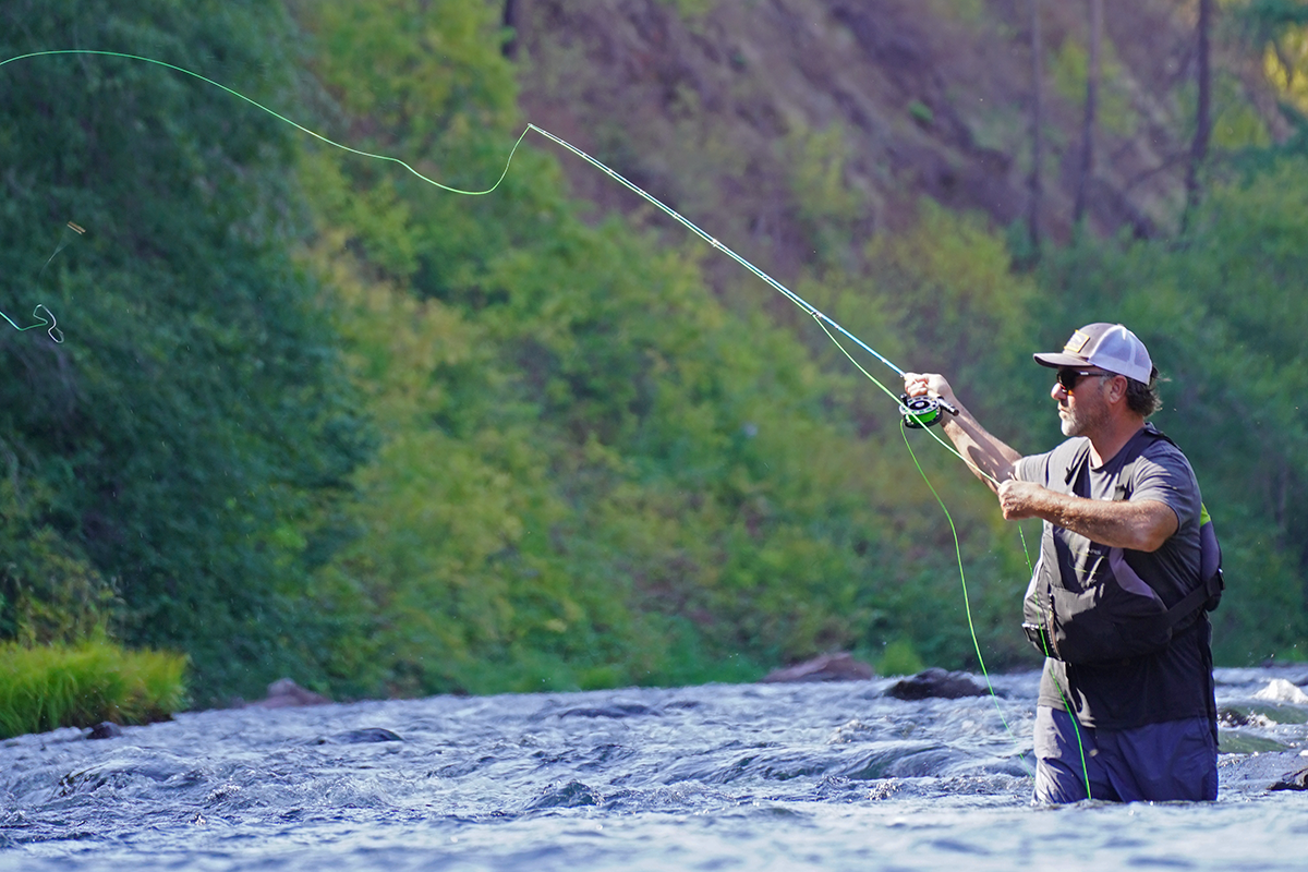 A man wearing sunglasses, a hat, and outdoor clothing, stands waist-deep in a river while fishing with a rod.