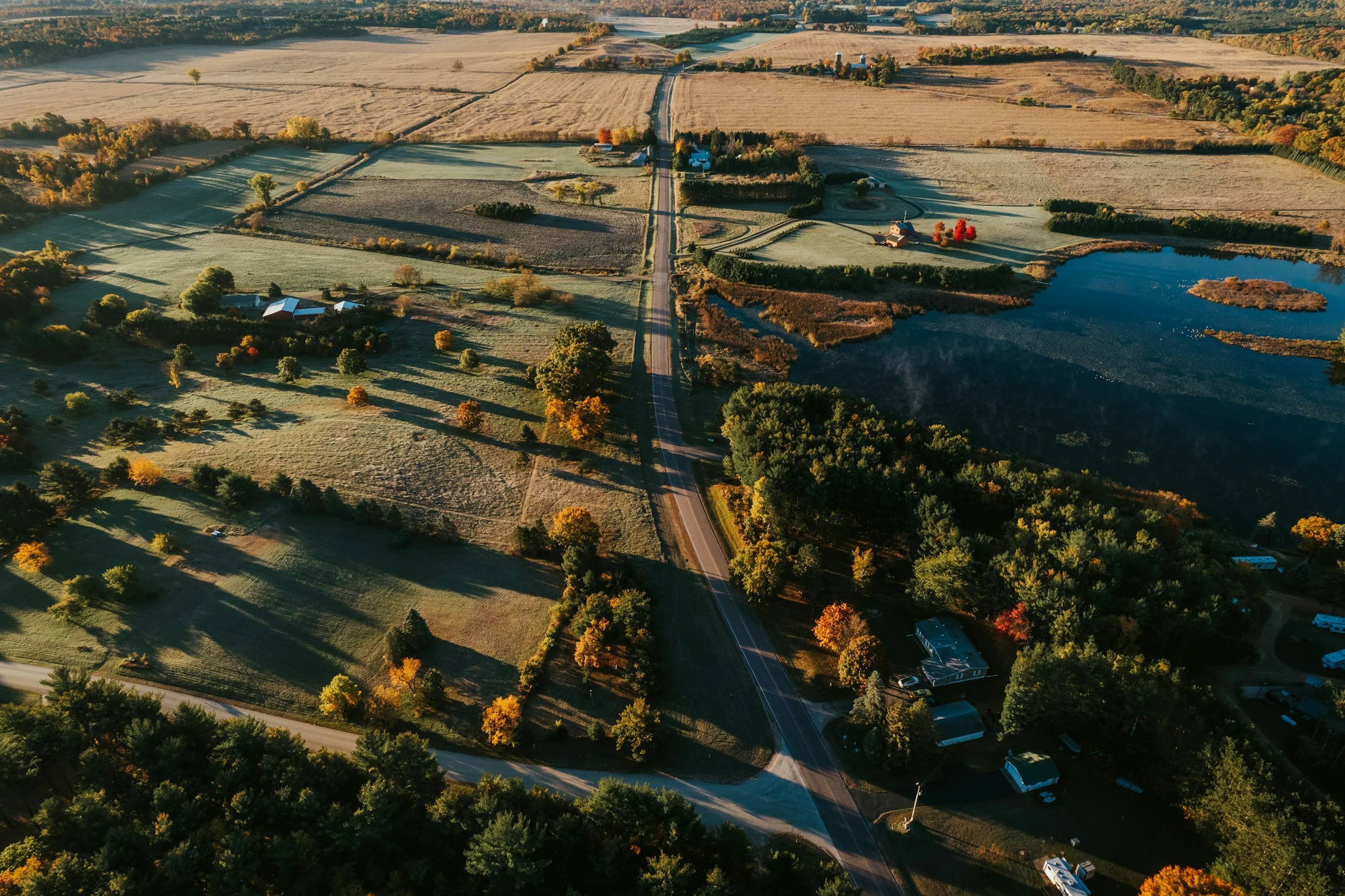 Aerial view of a rural landscape with open fields, a pond, scattered trees, houses, and a straight road running through the countryside during autumn.