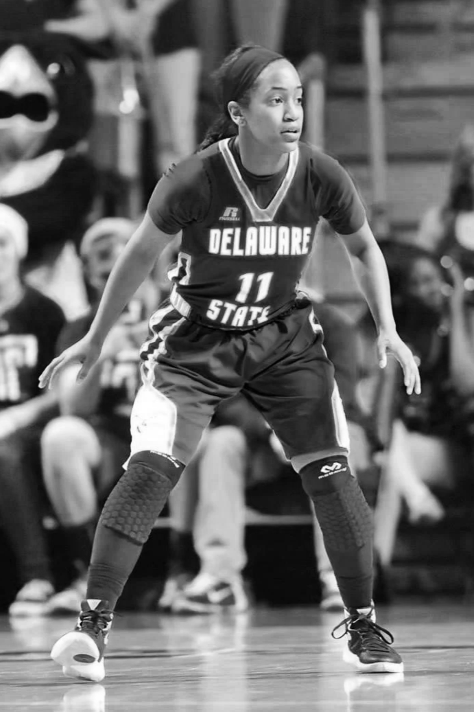 A female volleyball player in a Delaware State jersey, number 11, standing in a defensive stance on the court during a game.