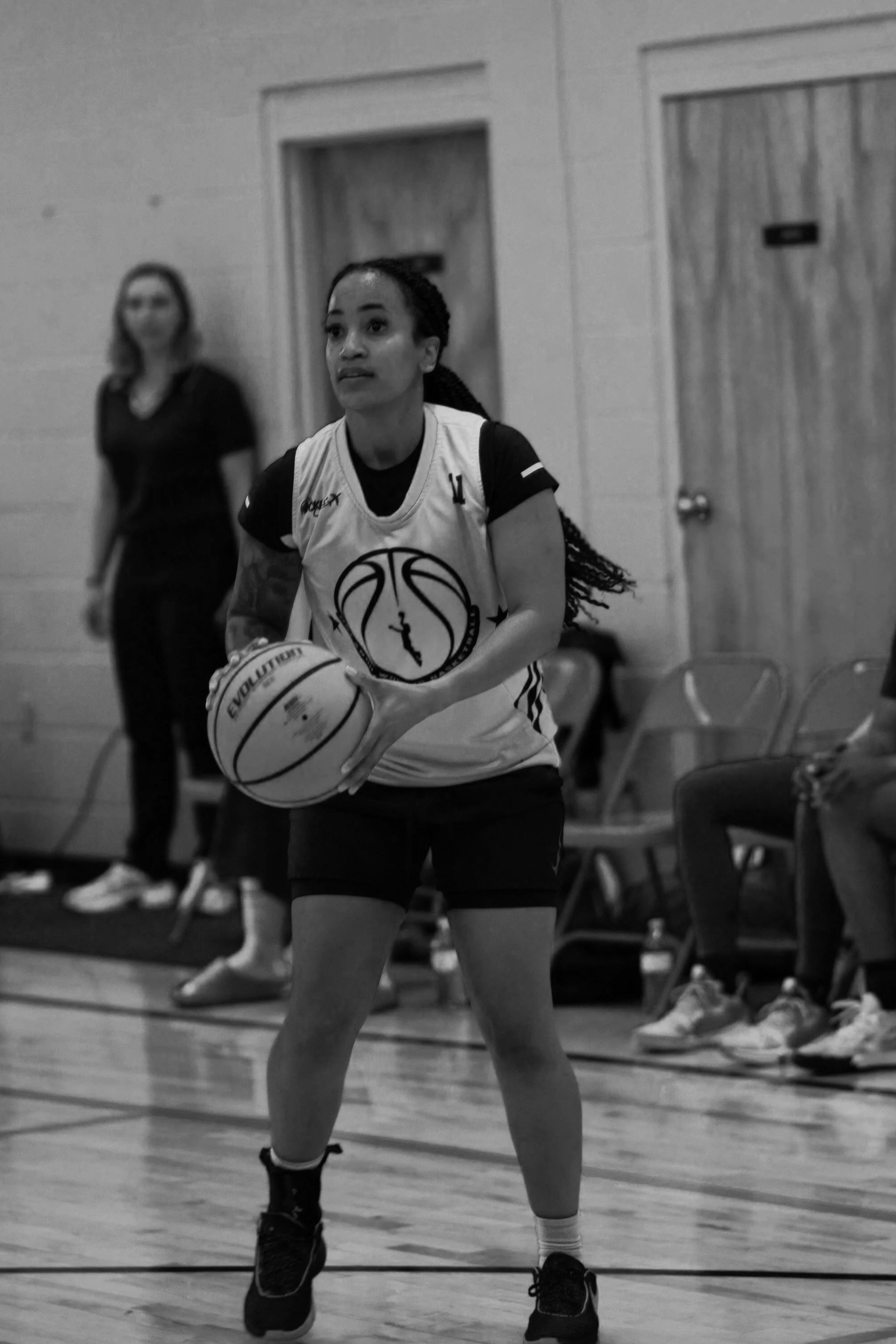 A woman in sports attire prepares to shoot a basketball on an indoor court, with another person standing in the background.