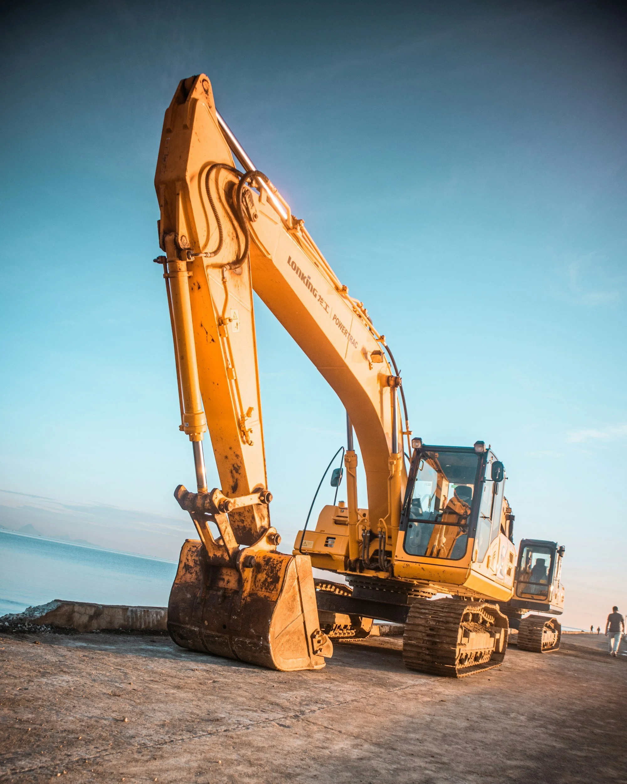 A yellow excavator on a sandy beach near water, with a person walking in the distance during sunset or sunrise.