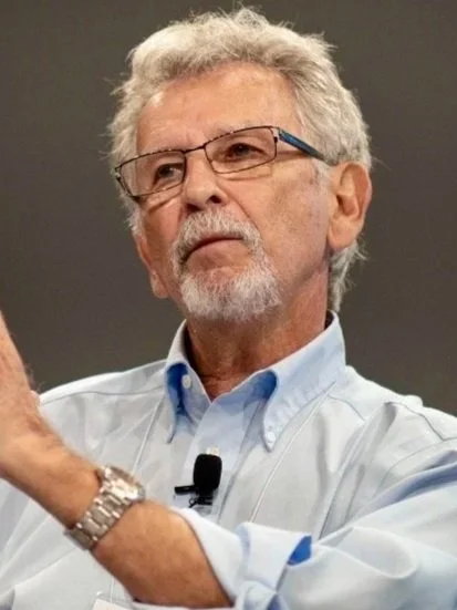 An older man with gray hair, glasses, and a beard, wearing a light blue shirt, appears to be speaking or presenting in an indoor setting.