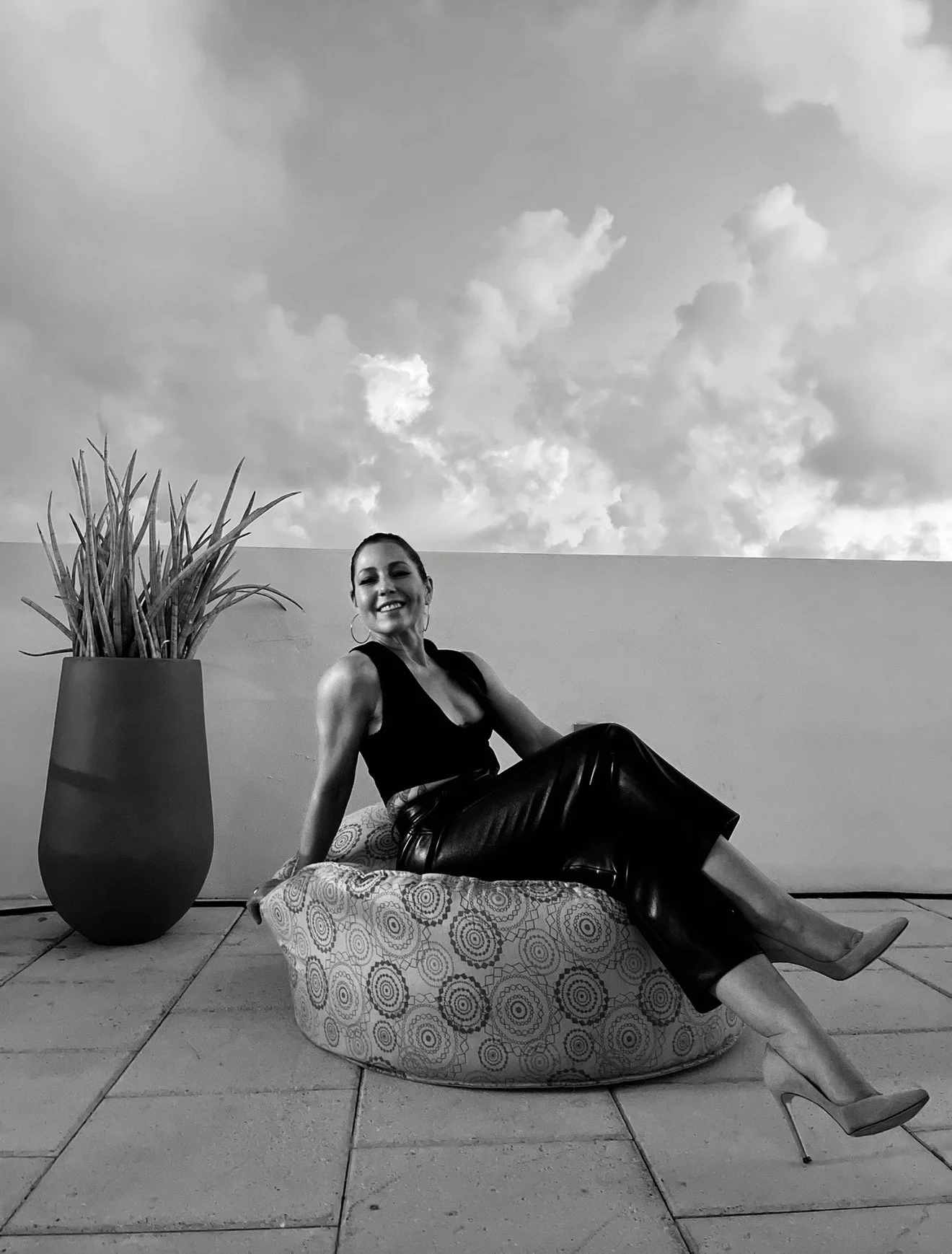 A woman wearing a sleeveless top, leather pants, and high heels, sitting on a patterned cushioned seat outdoors next to a large potted plant, smiling at the camera with a cloudy sky in the background.