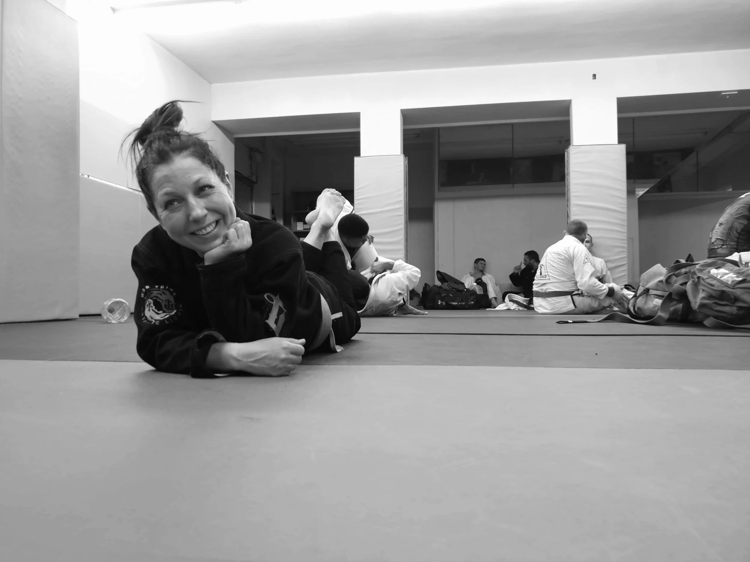 A woman in a martial arts gi lying on a mat, smiling, in a training facility with others practicing martial arts in the background.