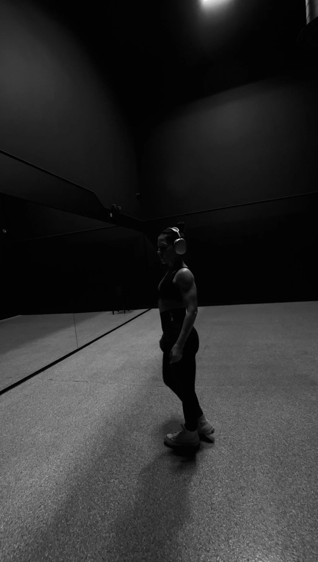 A person standing on a gym floor, wearing headphones, facing a mirror in a dimly lit workout room.