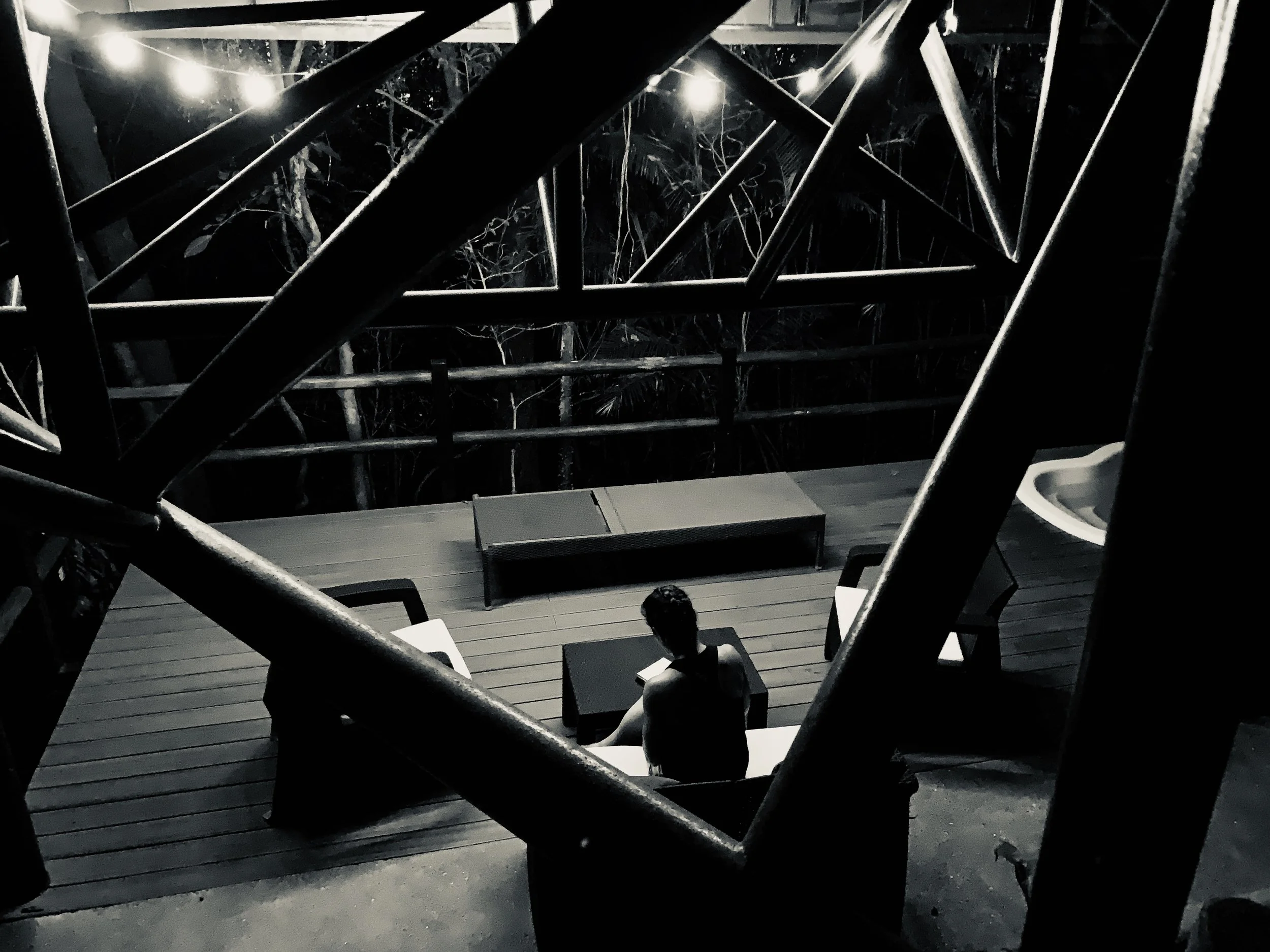 Nighttime view from a balcony with a person sitting on a chair, surrounded by a metal railing and trees in the background, illuminated by outdoor lights.