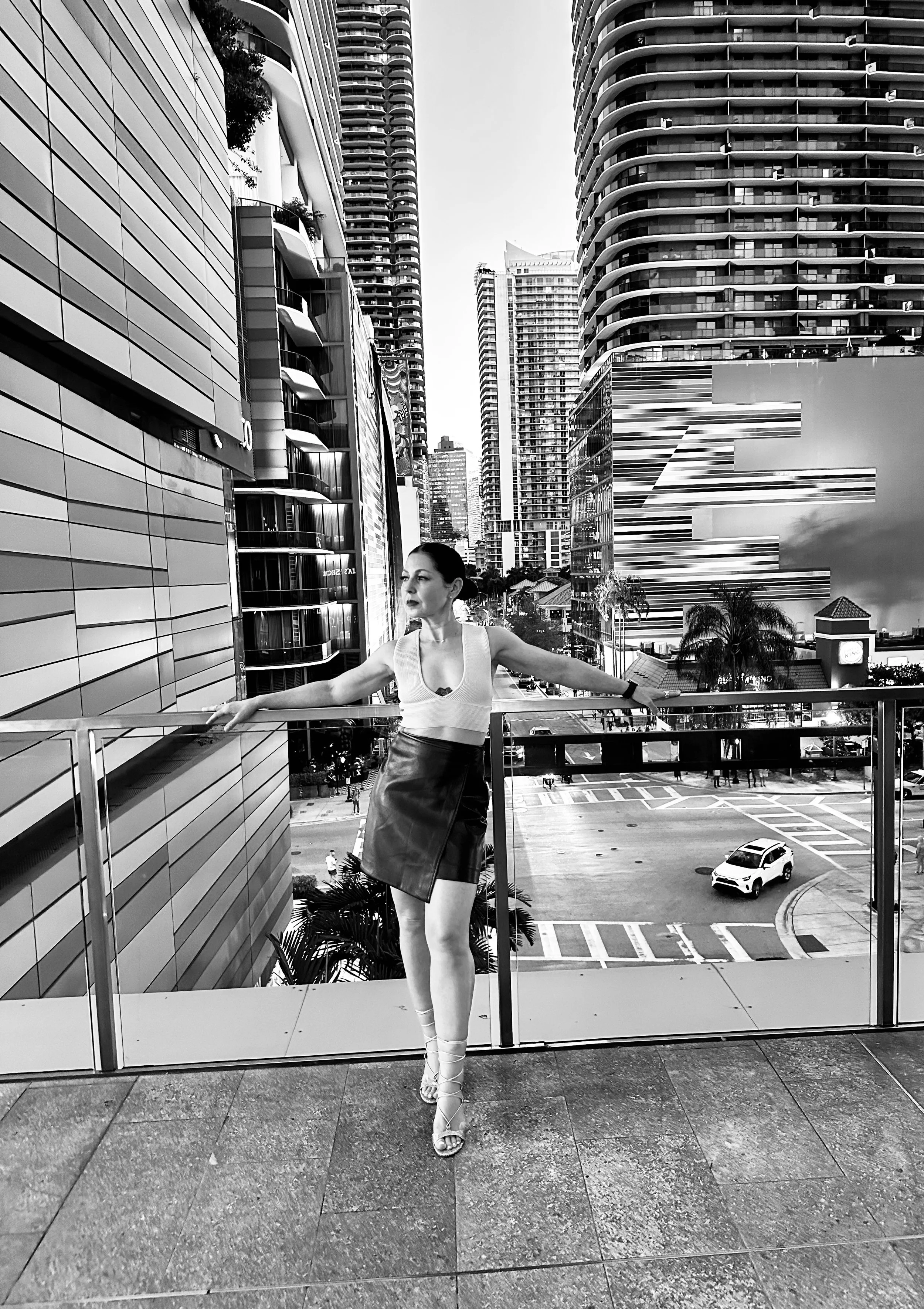 A woman standing on a balcony in an urban area with tall buildings and a parking lot visible in the background.