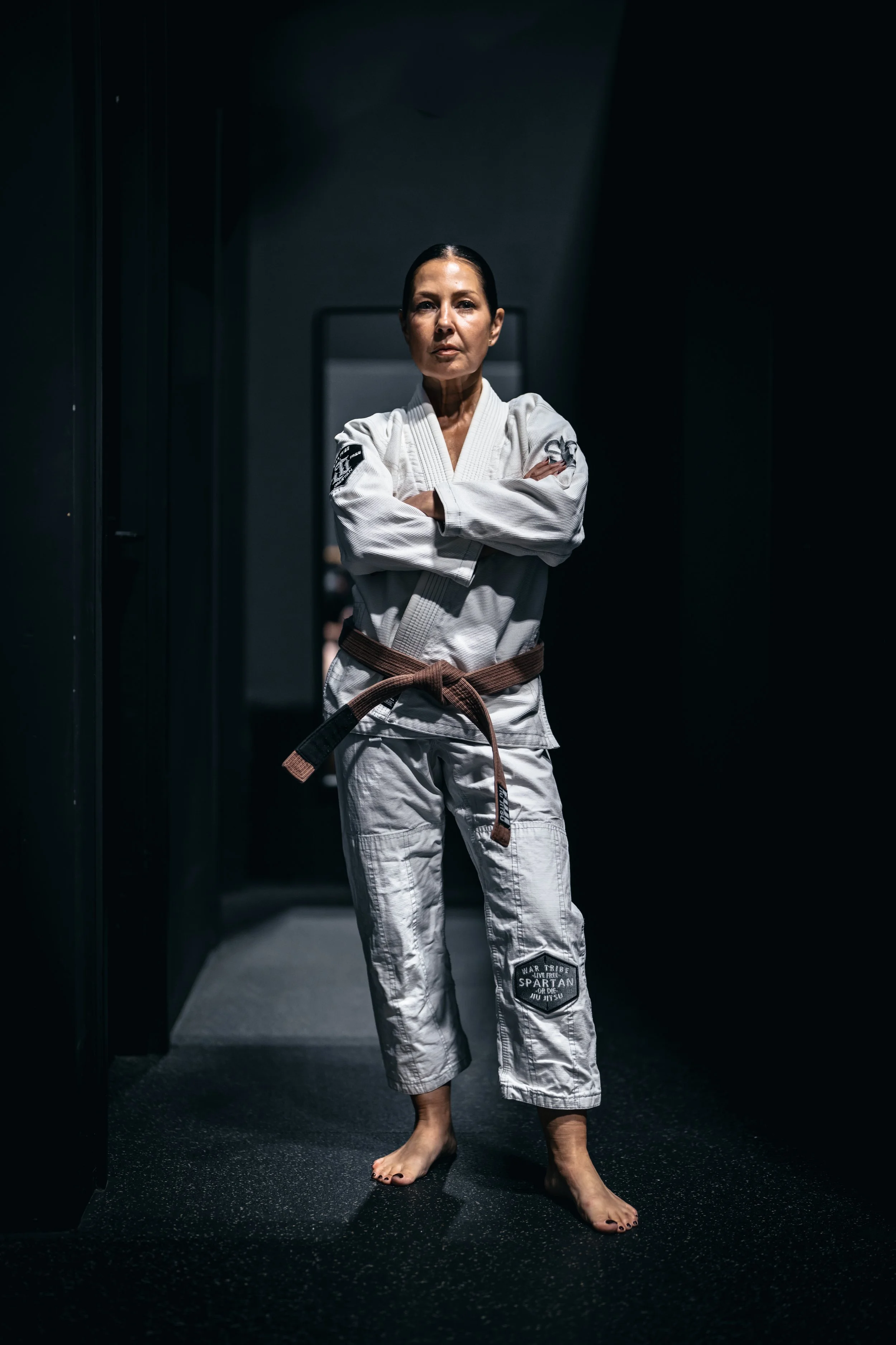 A woman standing barefoot in a martial arts gi with a brown belt, arms crossed, in a dimly lit hallway.