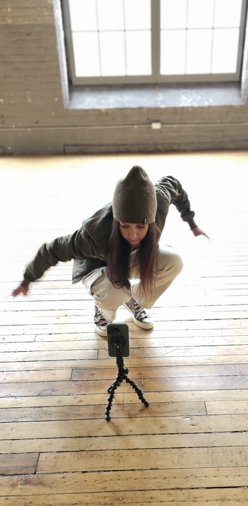 Person wearing a gray beanie, black jacket, and beige pants crouching down in front of a camera on a small tripod, on a wooden floor with a window in the background.
