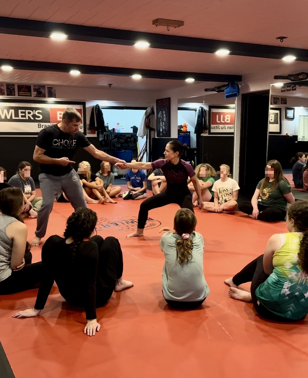 A martial arts class with children sitting on a red mat, watching an instructor and a woman practicing a self-defense move. The instructor is holding the woman's arm as she demonstrates. The room has mirrors, posters, and a sign in the background.