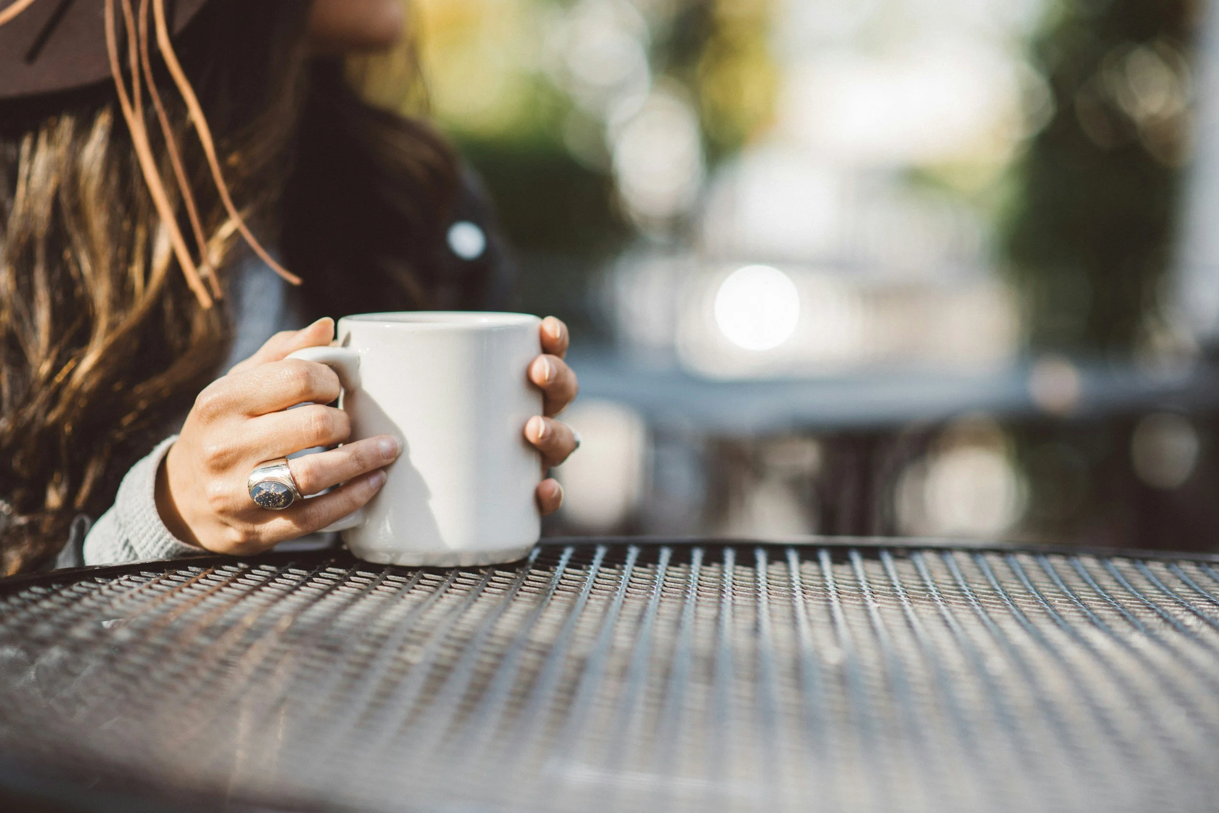 A person holding a white mug on a metal table outdoors with blurred greenery in the background.